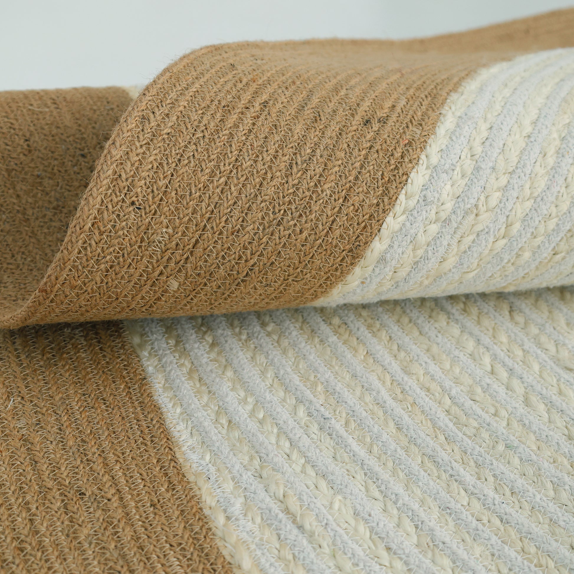 A close-up of the Home Decor Round Jute Rug - BR 089 shows its tan and off-white stripes, highlighting the fiber texture and detail as the rug is partially rolled.