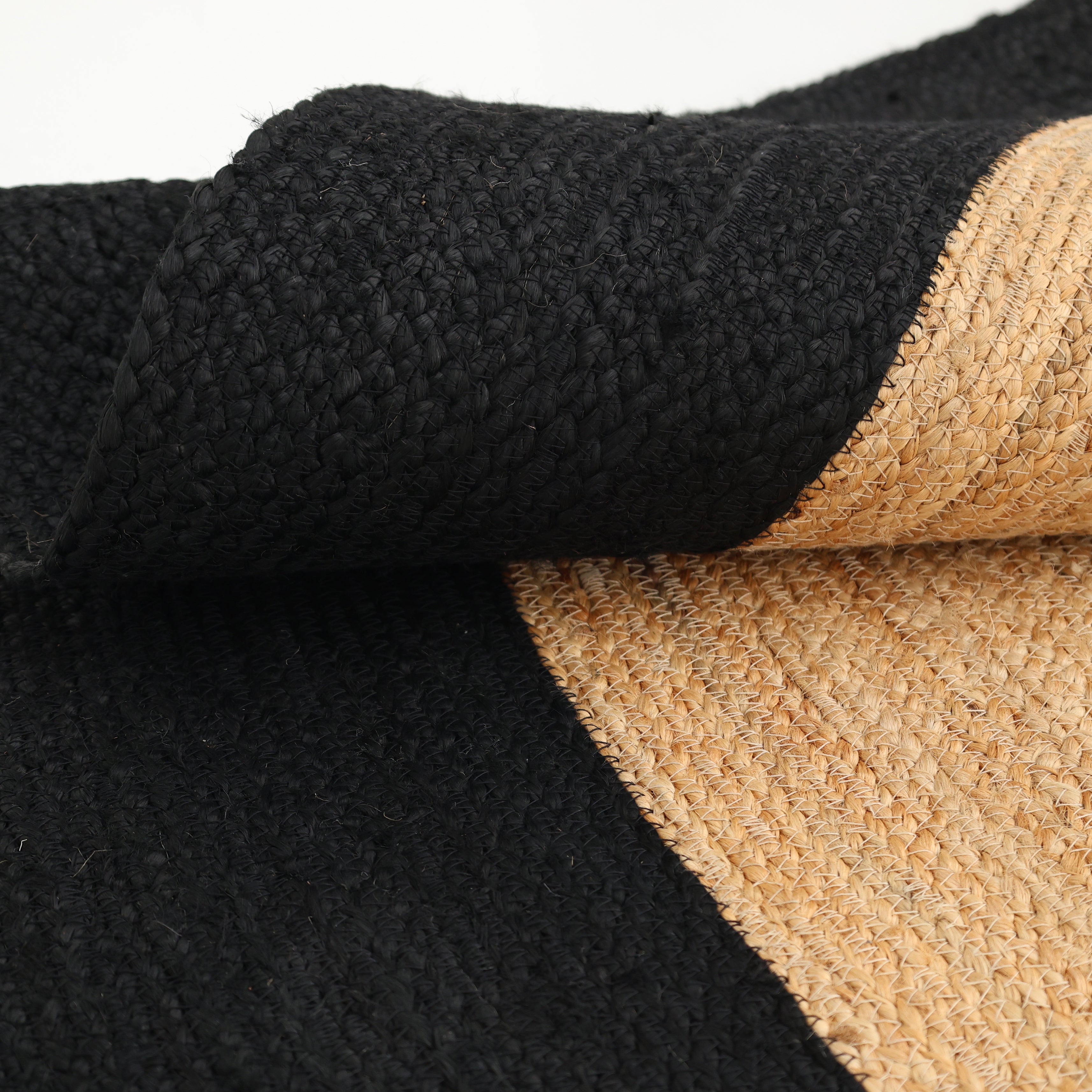 A close-up of the RAMSHA HOME Braided Rug Jute carpet BR-003, featuring natural beige and black fibers with a distinctive black border, partially folded to display its texture and quality craftsmanship.