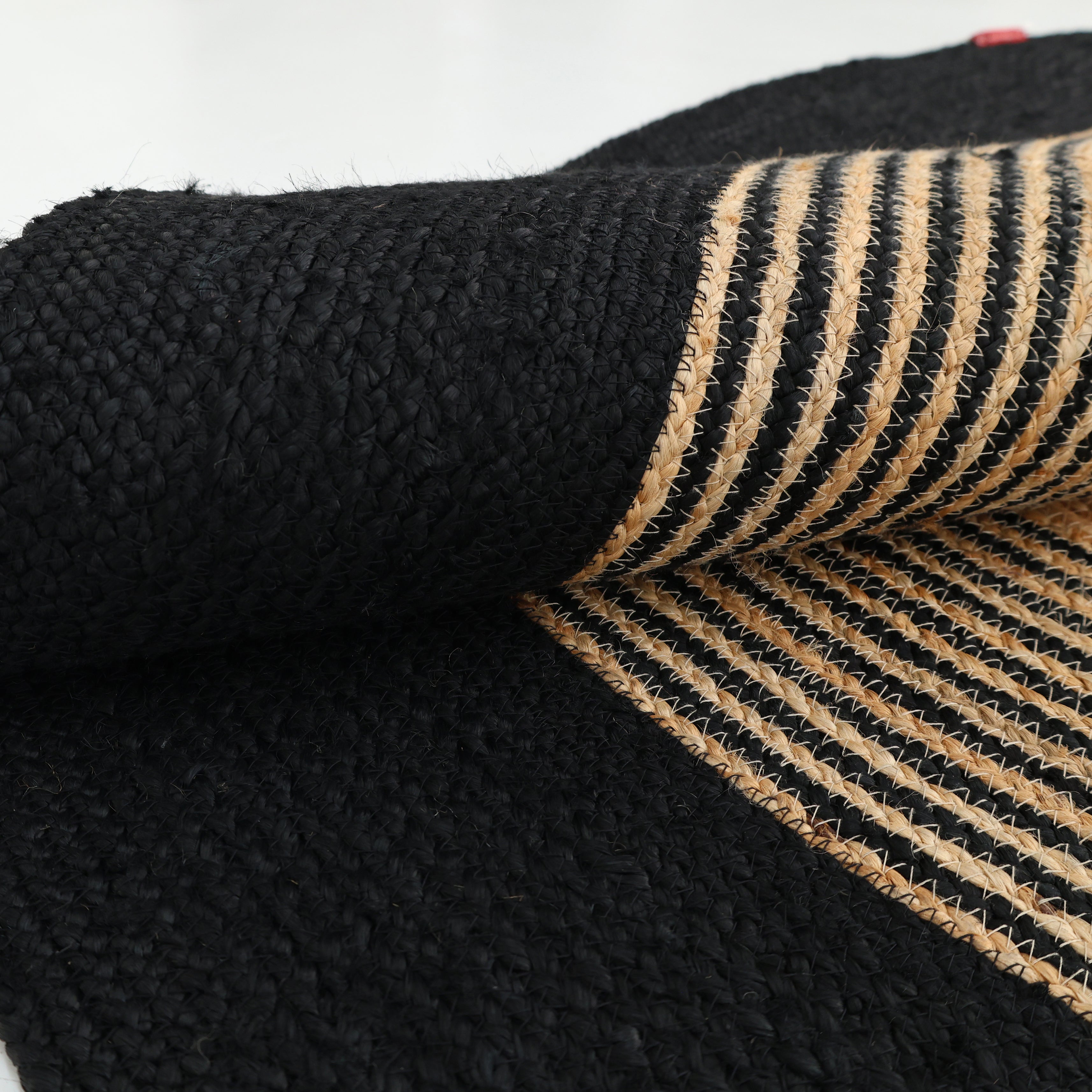 A close-up of RAMSHA HOME’s Luxurious Handmade Braided Jute Round Rug - BR 028, partly rolled up to display its striped underside and solid black top, elegantly highlights the timeless style of a black border rug.