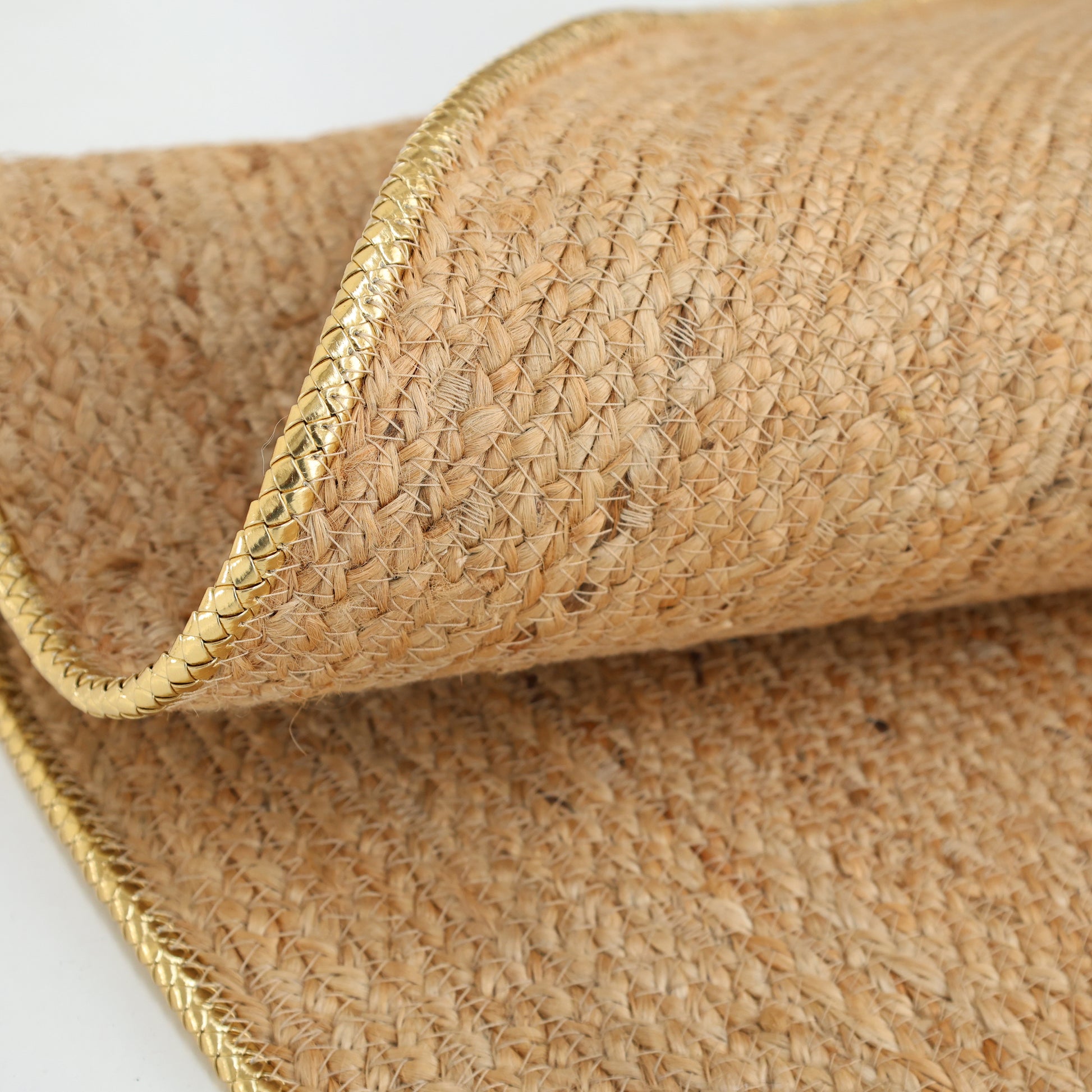 A close-up of the RAMSHA HOME Handmade Natural Color Braided Round Rug - BR 013, featuring gold-trimmed edges and shown partially folded on a white surface.