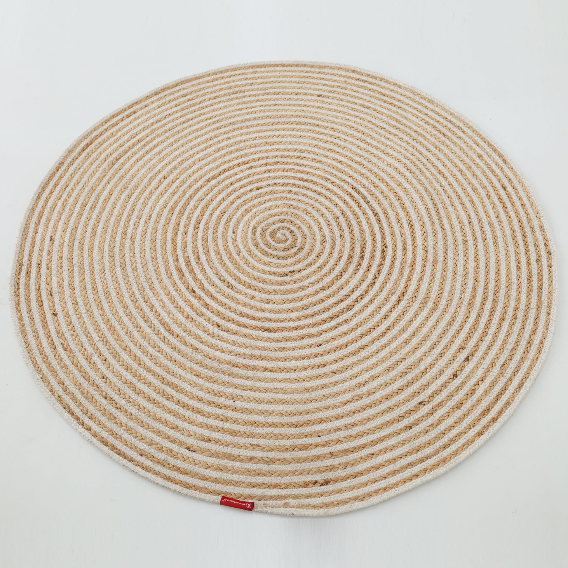The Handmade Natural & White Color Braided Jute Round Rug - BR 012, featuring a spiral pattern in beige and off-white, is shown on a white background with a small red tag attached to its edge.