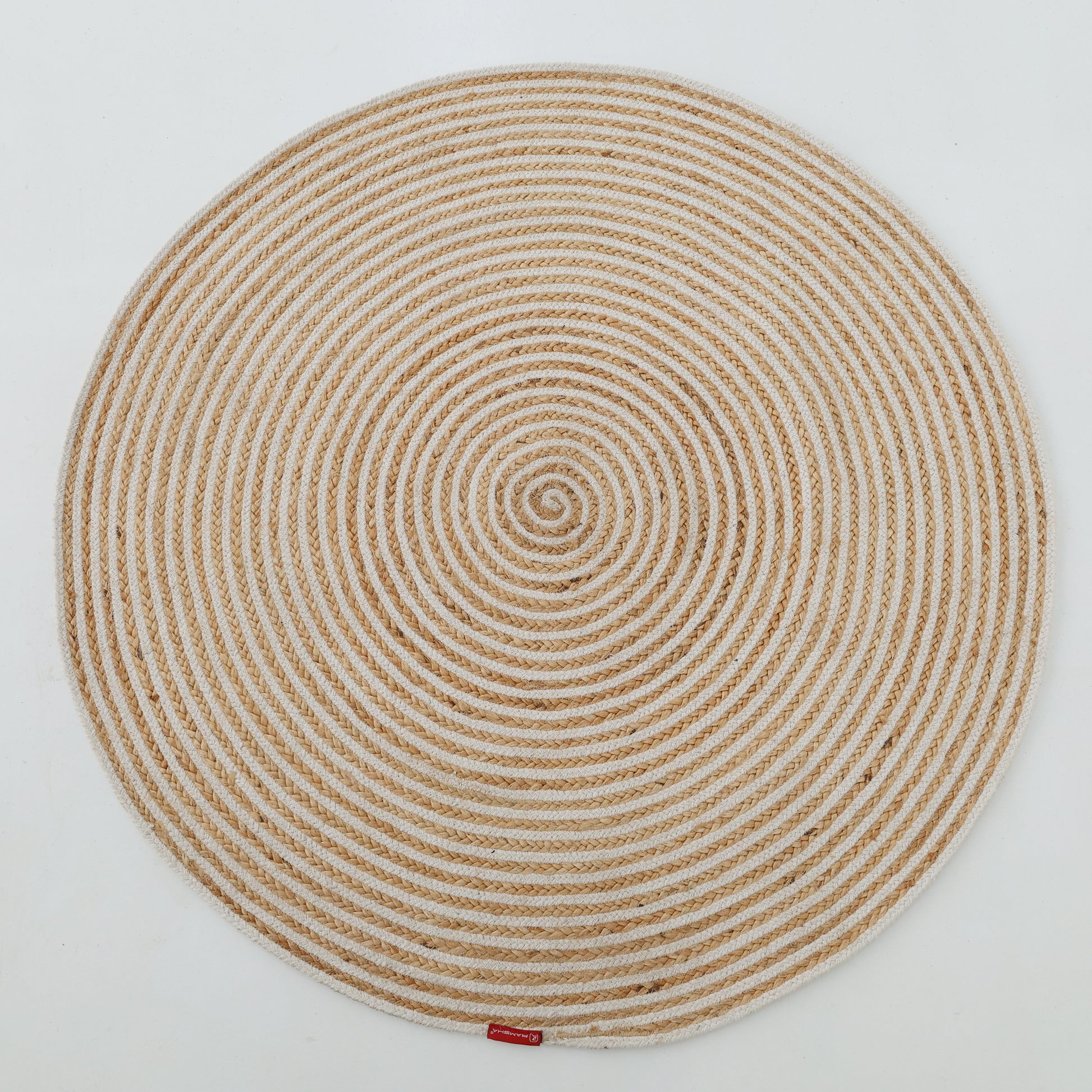 The Handmade Natural & White Color Braided Jute Round Rug - BR 012, featuring a spiral braided pattern in beige and white and a small red label near the edge, is displayed on a plain white background.