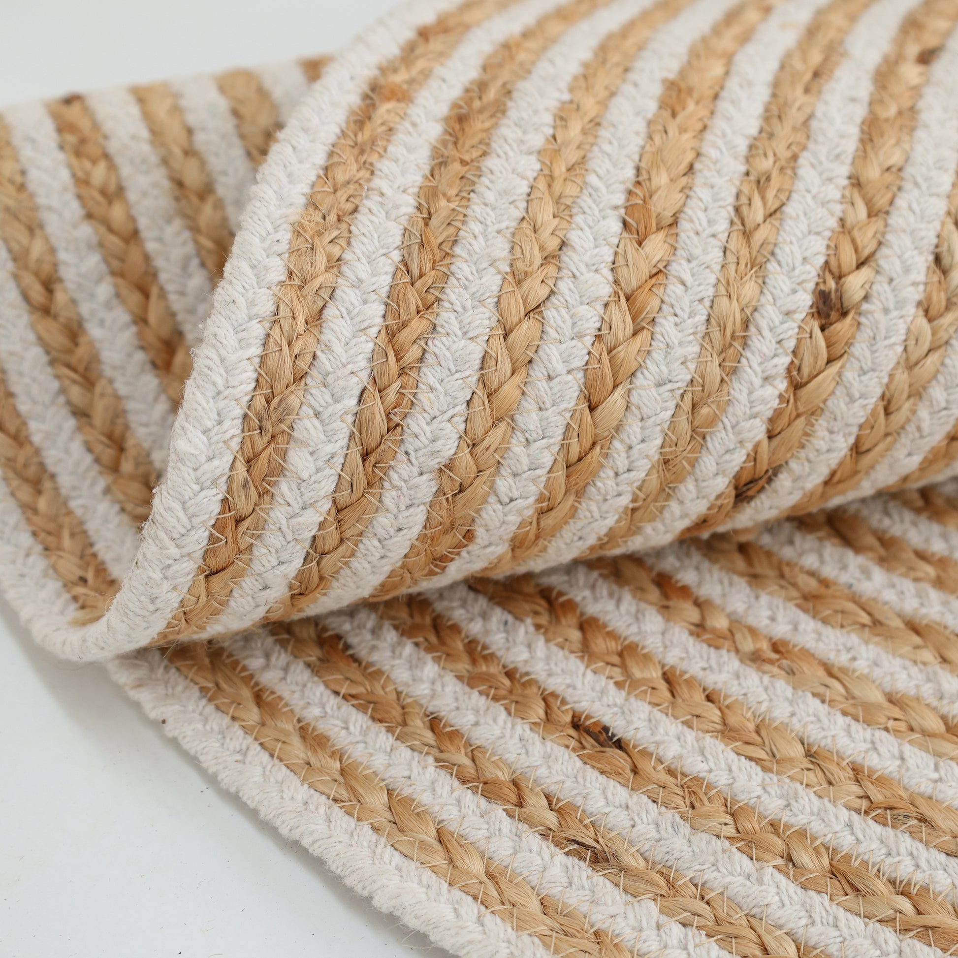 A close-up of the Handmade Natural & White Color Braided Jute Round Rug - BR 012, featuring alternating beige and white stripes, is partially rolled up on a white surface.