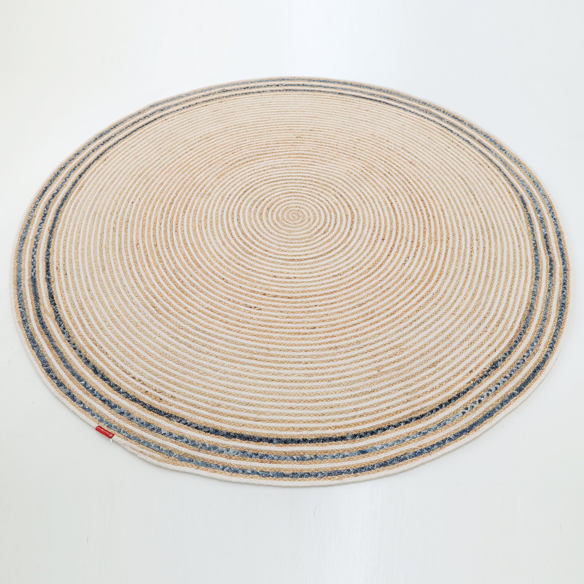 The RAMSHA HOME Creative Handcrafted Blue Border Braided Rug - BR 065 features concentric circular patterns in beige, gray, and white, accented with a blue border and a small red tag on the edge, stylishly placed on a white floor.