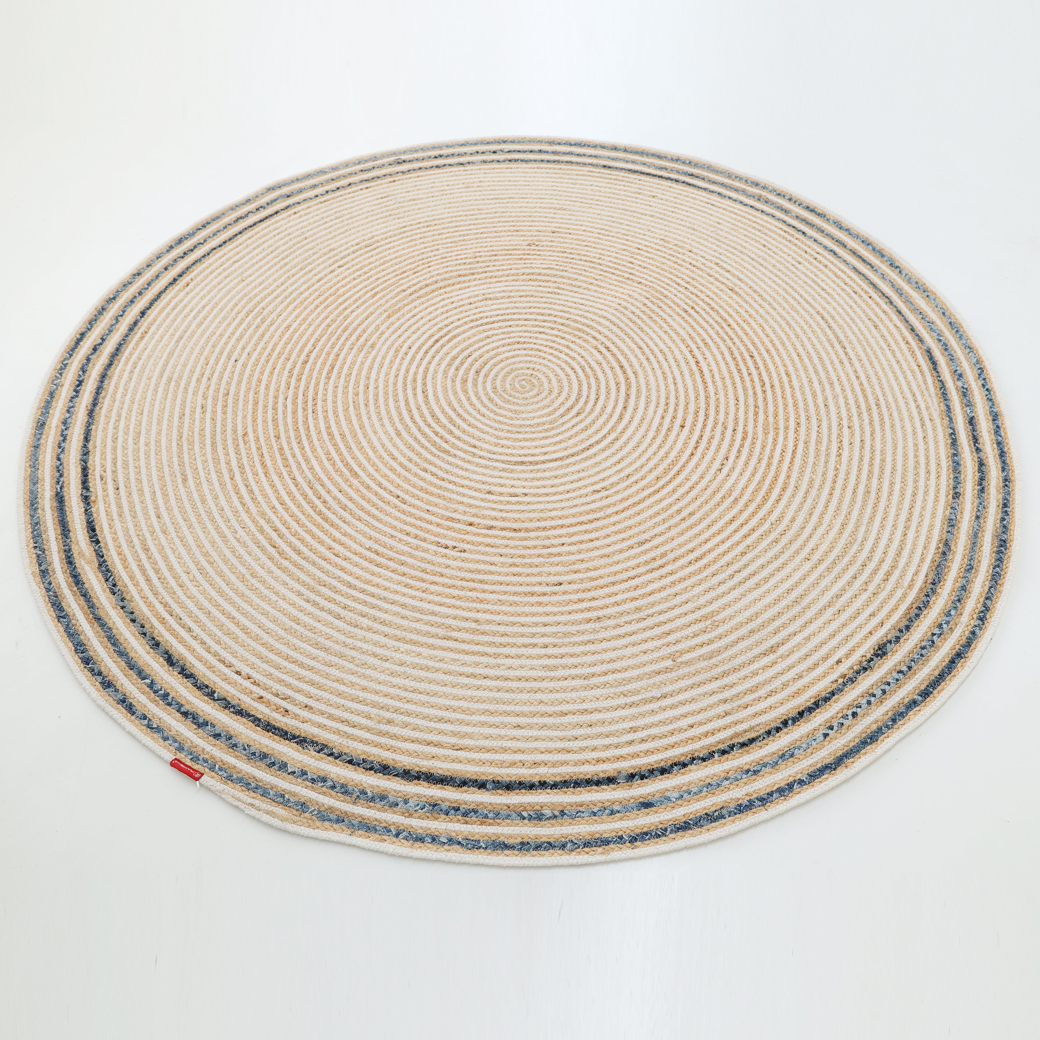 The RAMSHA HOME Creative Handcrafted Blue Border Braided Rug - BR 065 features concentric circular patterns in beige, gray, and white, accented with a blue border and a small red tag on the edge, stylishly placed on a white floor.