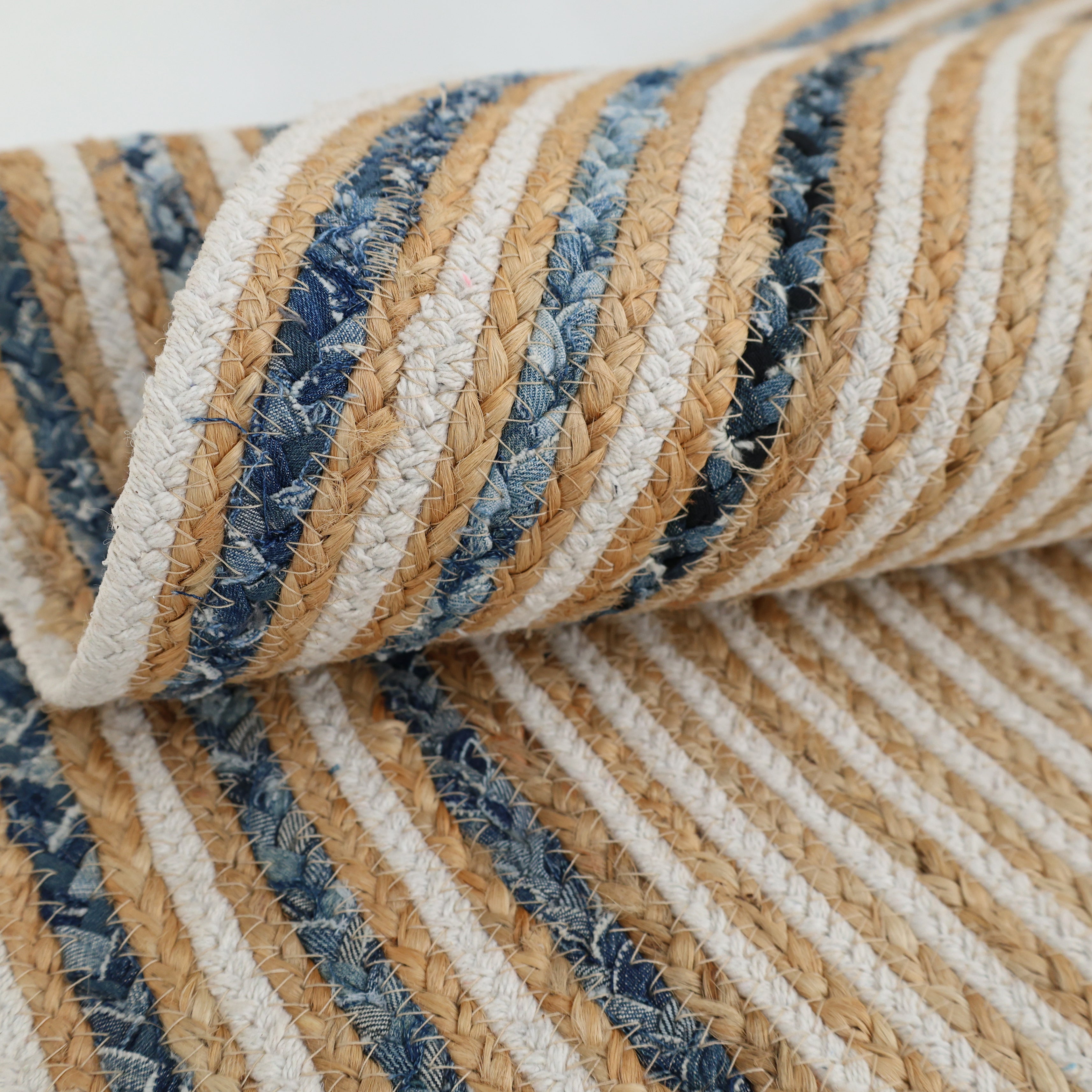 A close-up of the RAMSHA HOME Creative Handcrafted Blue Border Braided Rug - BR 065 shows its coarse, woven texture and alternating beige, white, and blue stripes. The round rug, partially rolled up, highlights its detailed braided design.