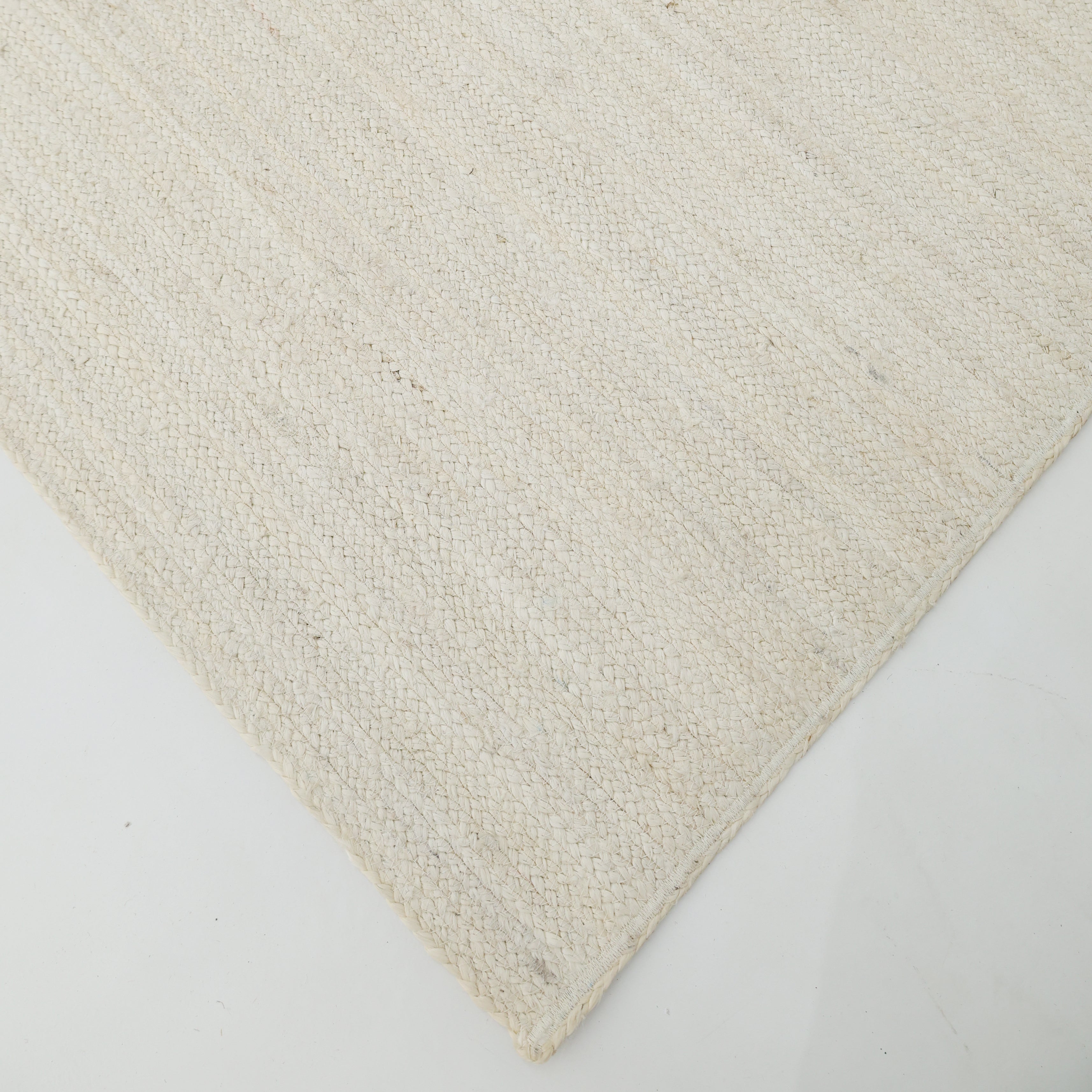 A close-up of the Amazing Handmade Bleach Jute Braided Rug - BR 019 by RAMSHA HOME shows its textured, light beige surface and subtle pattern in a corner view on a white floor.