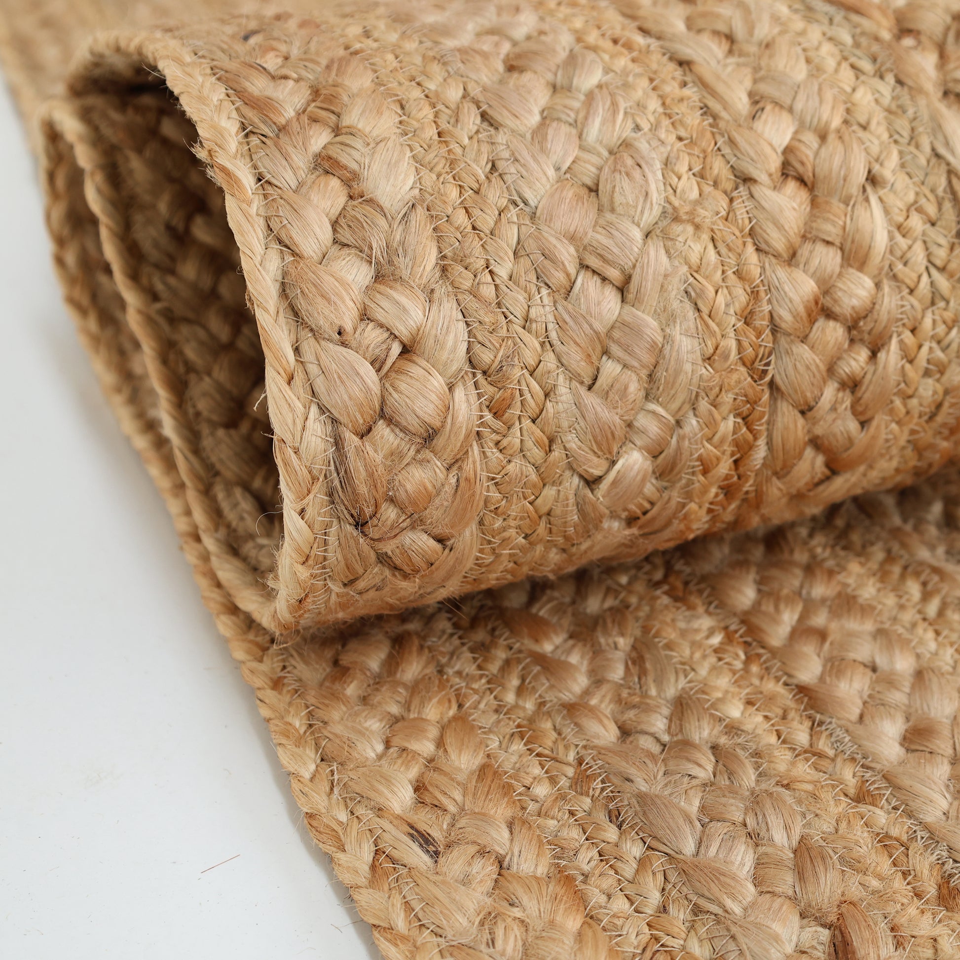A close-up of the RAMSHA HOME Braided Jute Natural Color Handmade Rug - BR 021, showcasing its rolled, natural fiber texture and light brown hue on a white surface.