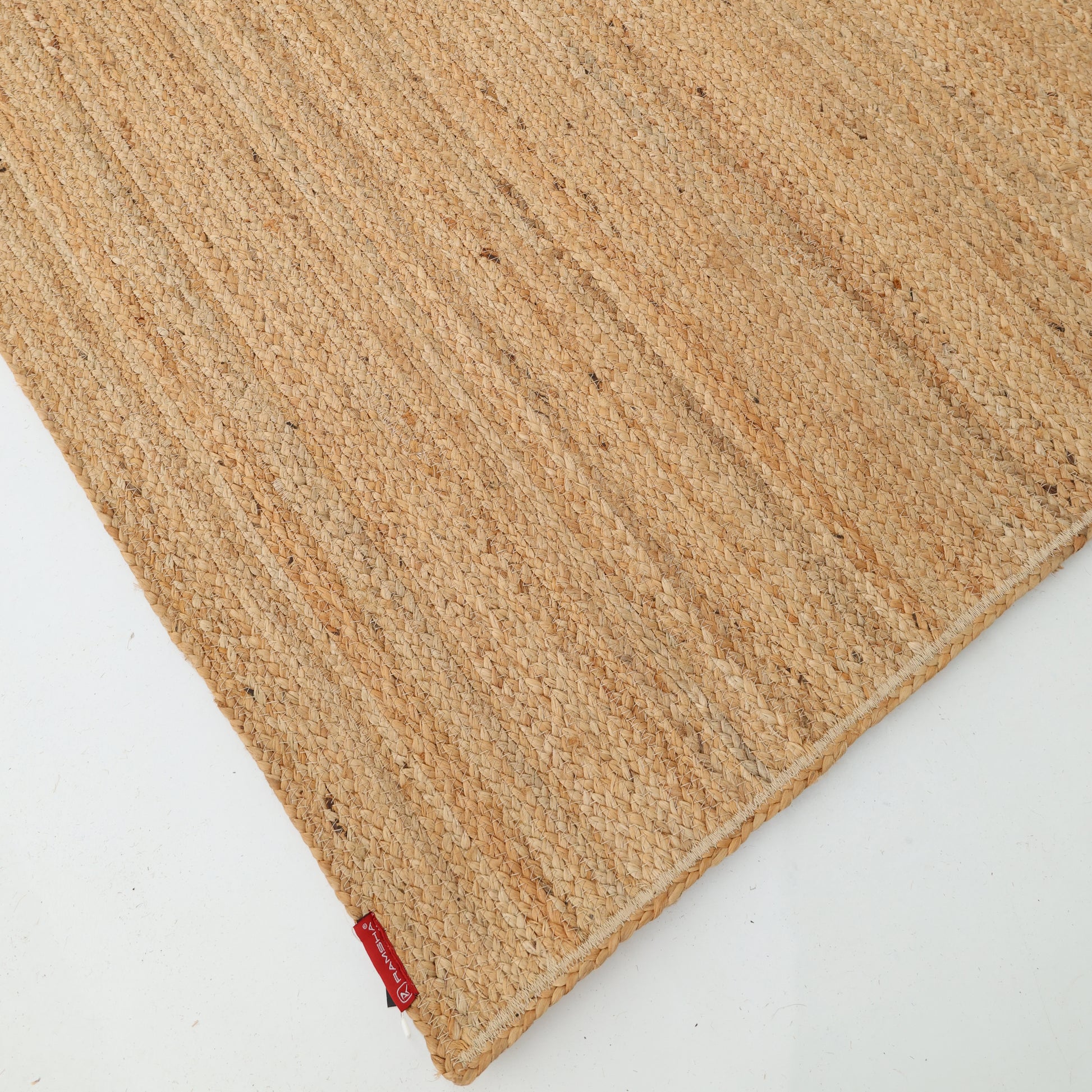 A close-up of RAMSHA HOME's Handmade Natural Color Braided Jute Rug - BR 018 on a white surface, showcasing its textured pattern and a small red fabric tag in one corner.