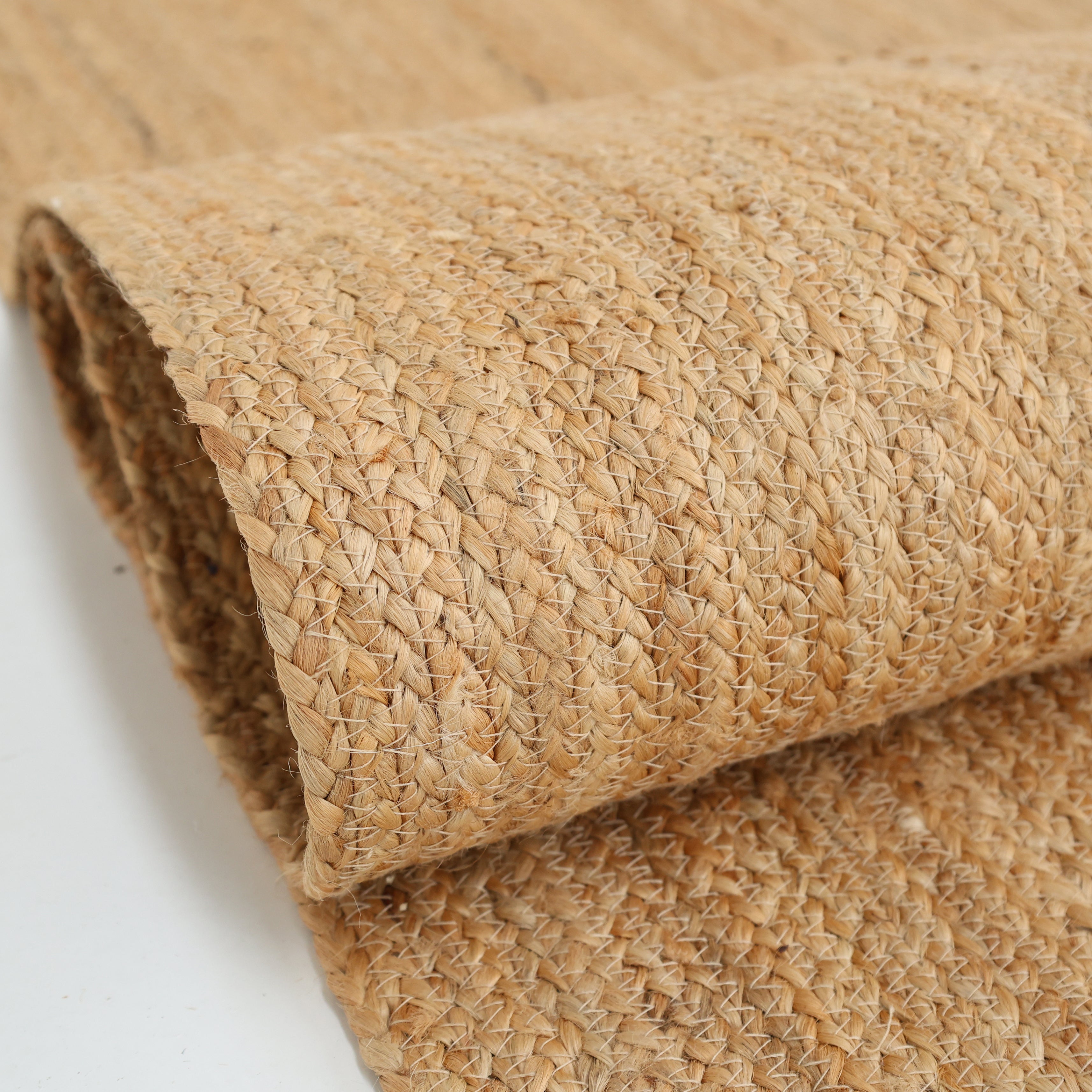 A close-up of the RAMSHA HOME Handmade Natural Color Braided Jute Rug - BR 018 shows its textured pattern and tan natural fibers, emphasizing this rug’s handcrafted beauty against a white background.