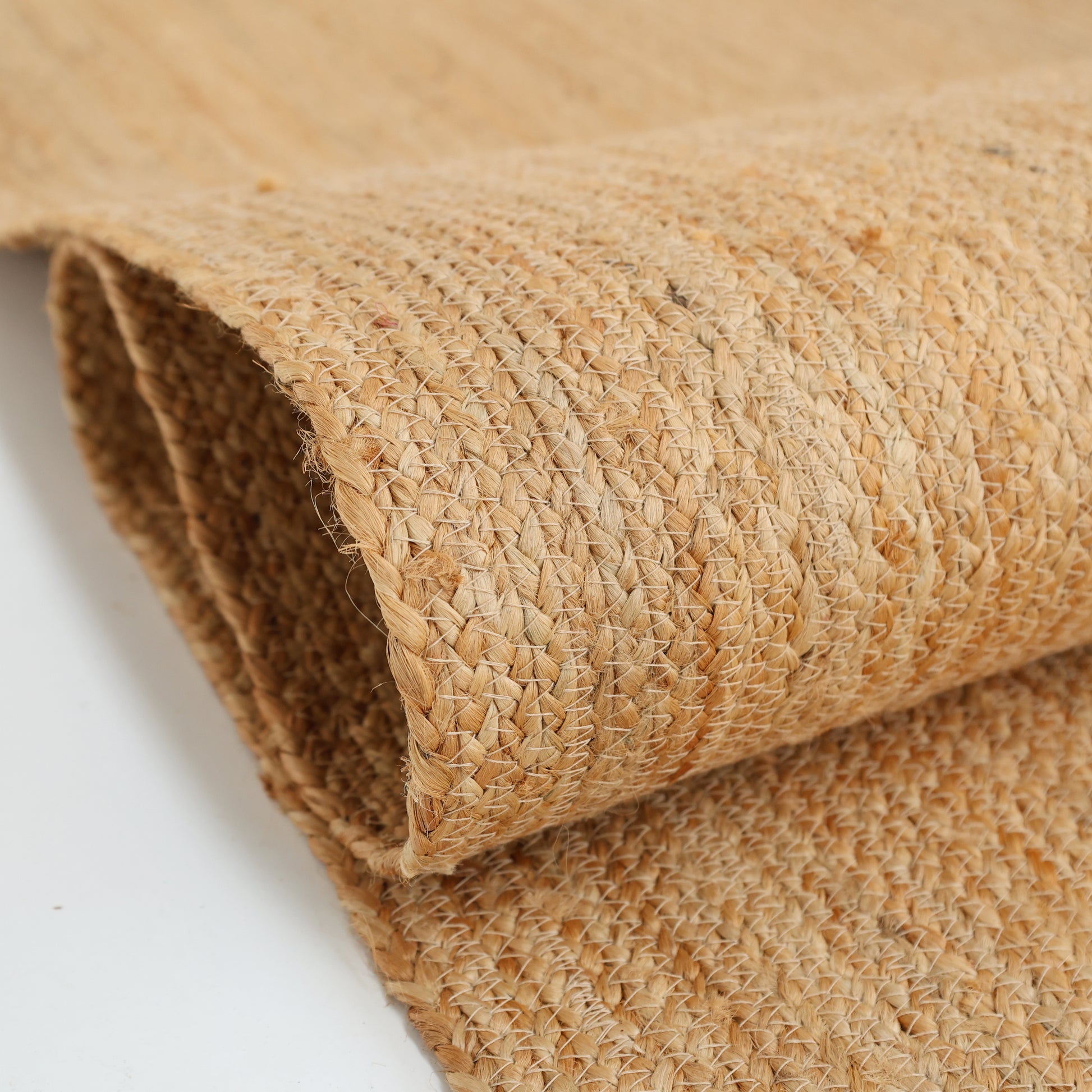 A close-up of the Handmade Natural Color Drawing Room Jute Floor Braided Rug - BR 046 highlights its coarse texture and light brown hue, crafted from natural jute fibers and displayed rolled up on a white surface.