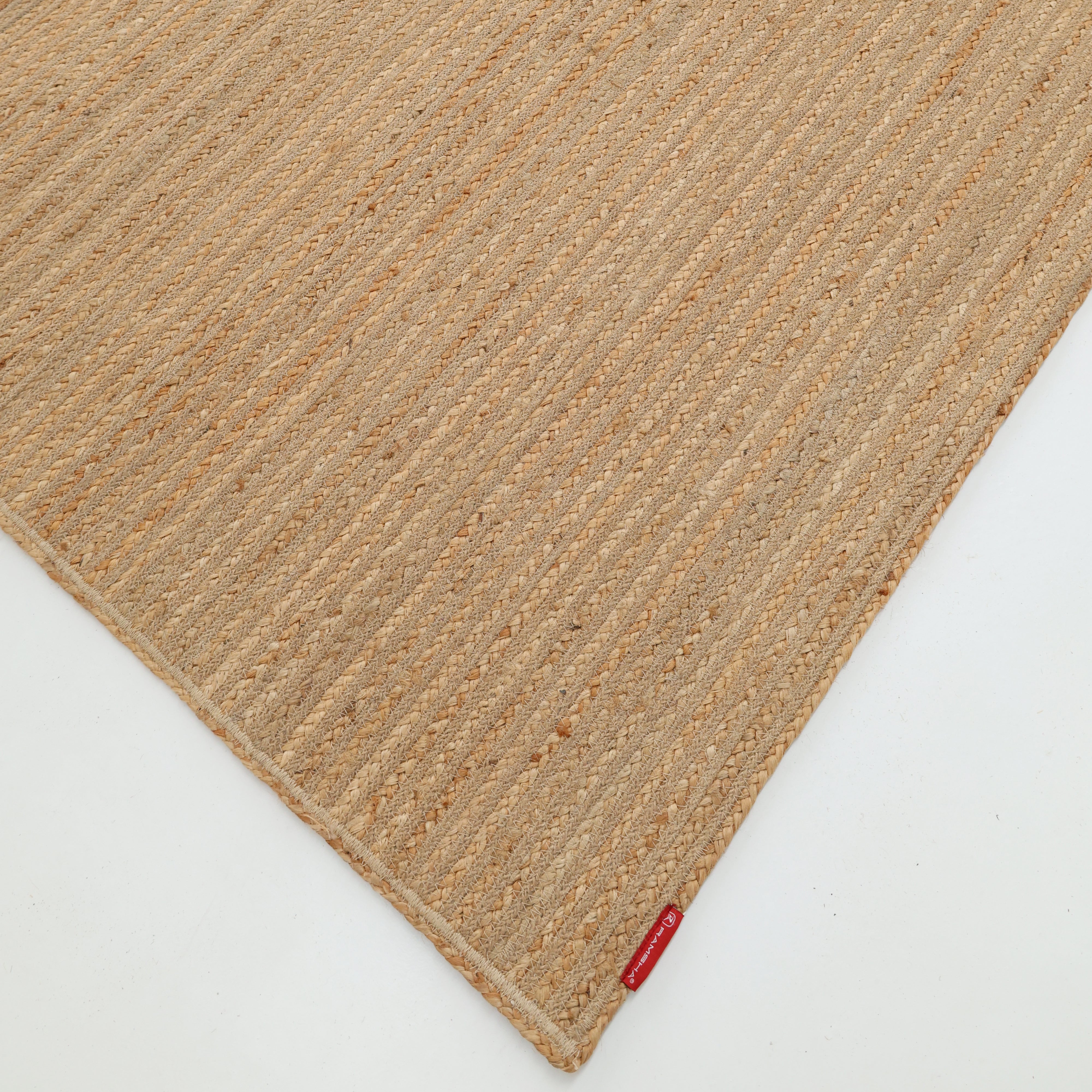 The Luxurious Handmade Jute Home Decor Floor Braided Rug - BR 087 by RAMSHA HOME is a beige rectangular rug with a subtle striped texture, shown angled on a white floor, featuring a small red fabric tag on one corner.