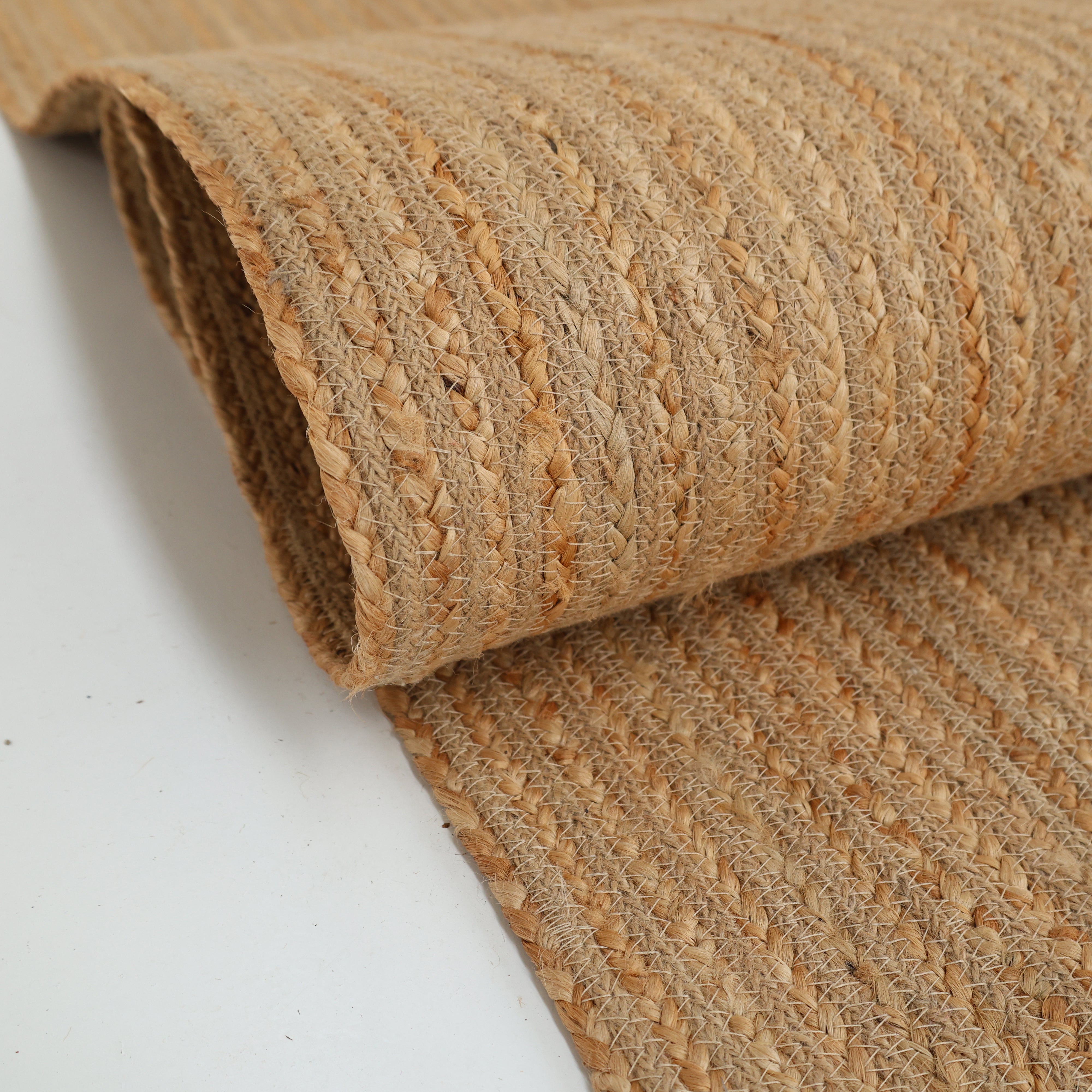 A close-up of the RAMSHA HOME Luxurious Handmade Jute Home Decor Floor Braided Rug - BR 087, showcasing its natural tan hue and textured braided patterns, neatly rolled and displayed on a white surface.