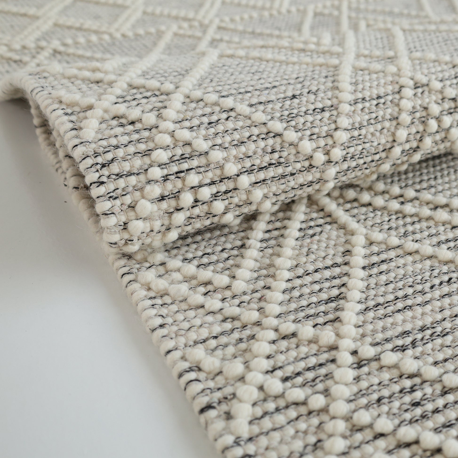 A close-up of the RAMSHA HOME Wool & Cotton Living Room Woven Rug SRN-019 shows its neutral tones, grey geometric tufted design, and subtle diamond pattern. The slight fold highlights the rug’s texture and substantial thickness.