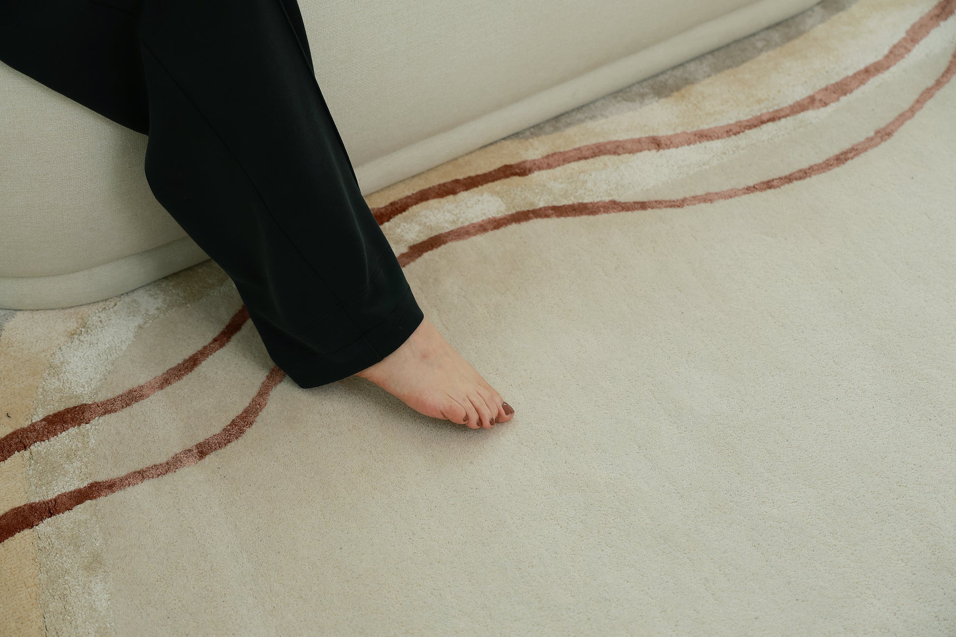 A person in black pants sits on a light sofa, one bare foot resting on the RAMSHA HOME Handmade Hand-Tufted Carpet HTWV-60, a cream rug adorned with curved red and beige lines.