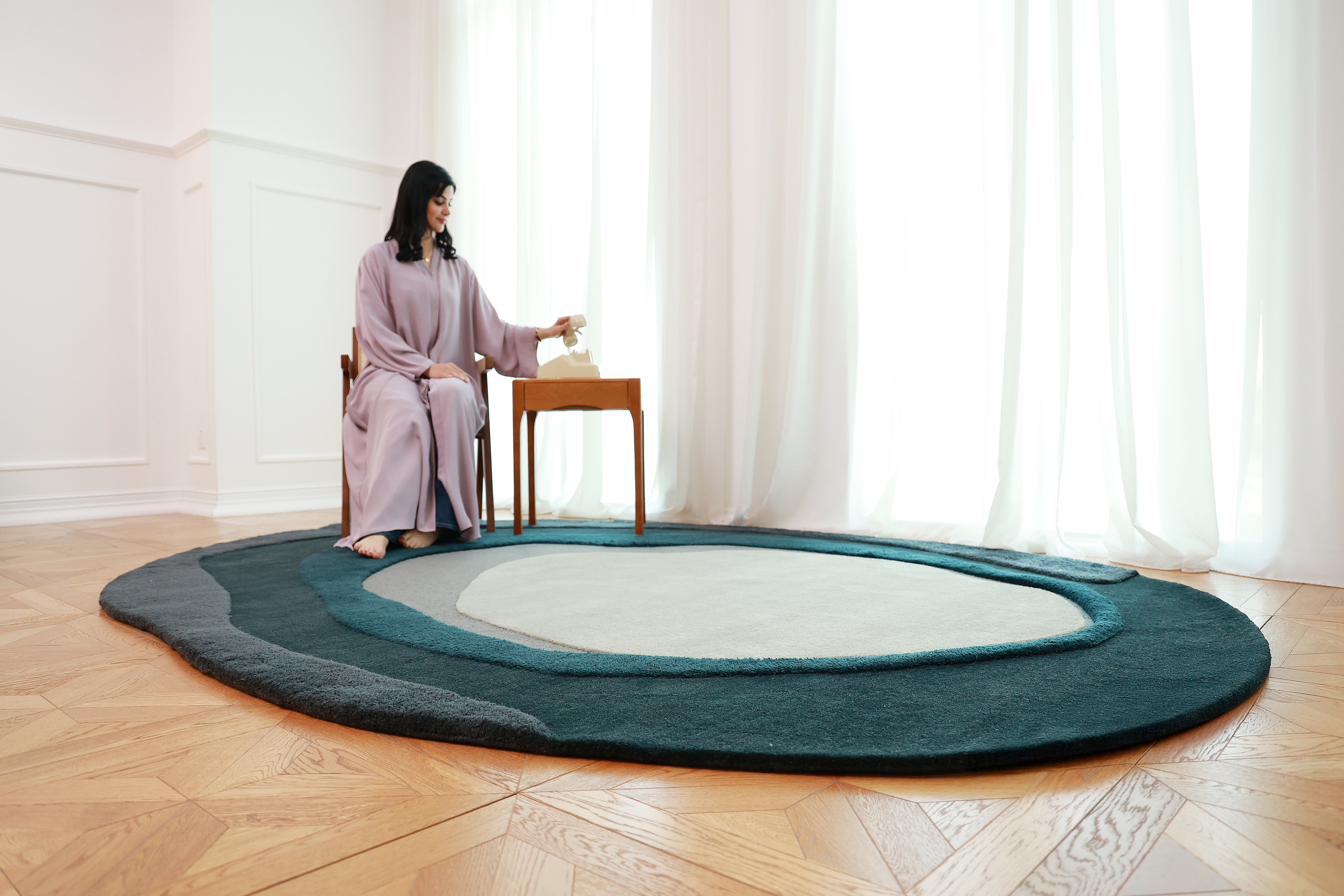 A woman in a light purple dress sits beside a small wooden table on the RAMSHA HOME Handmade Layered Abstract Shaped Rug HTW 131 (200 x 300 cm), featuring blue and gray tones, in a bright room with white walls and sheer curtains.