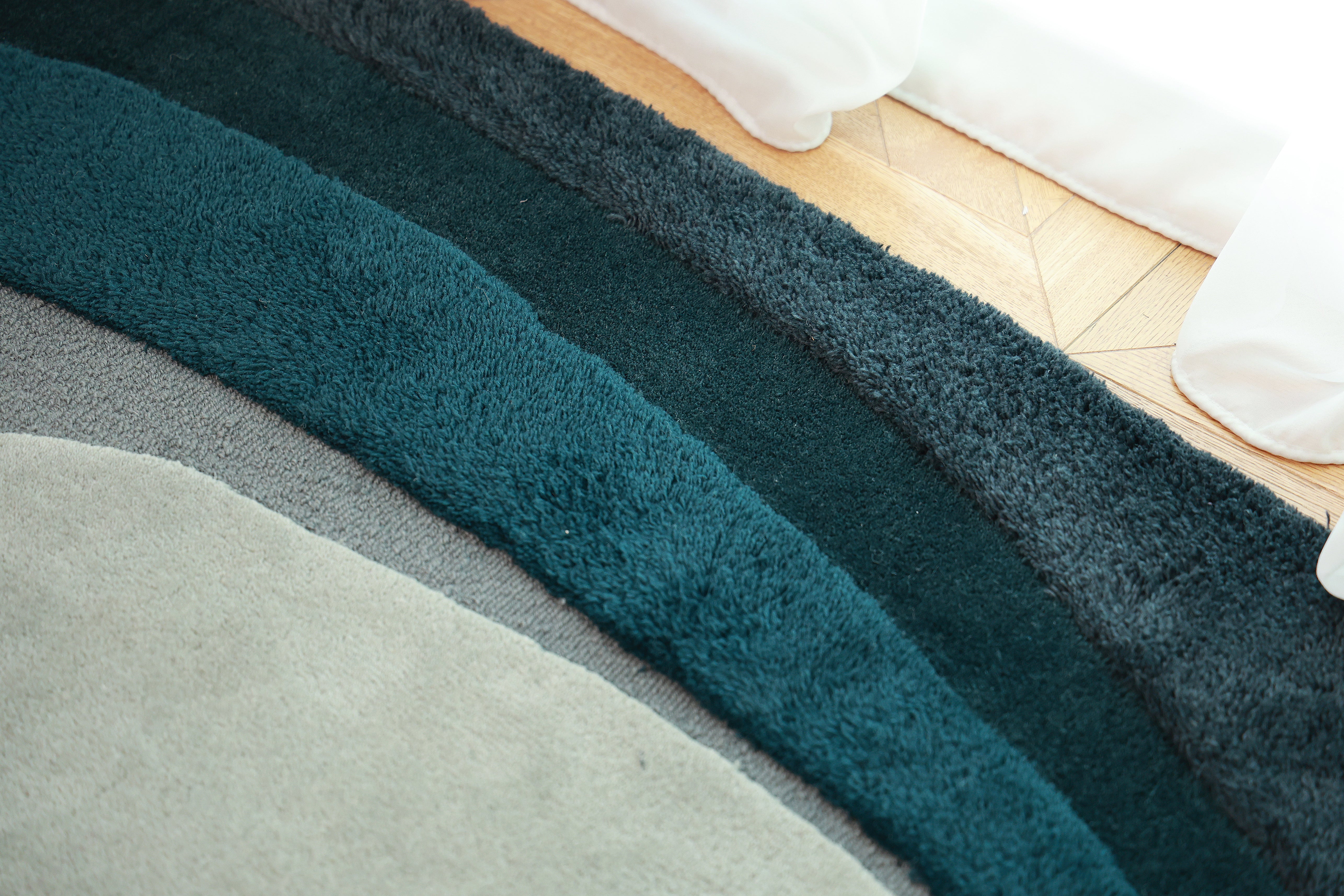 A close-up of the RAMSHA HOME Handmade Layered Abstract Shaped Rug HTW 131 (200 x 300 cm) showcases its thick, curved sections in gray, teal, and dark green on a wooden floor near white curtains and its distinctive irregular shape.