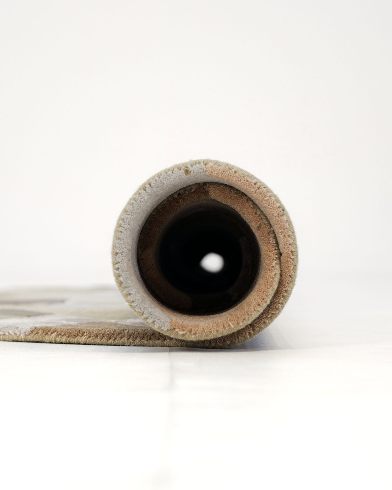 A rolled-up RAMSHA HOME Handmade Round Carpet For Home Decor 200 CM HTWV-47 in beige is lying on a white floor, viewed from the side and displaying its circular cross-section and hollow center against a plain, light background.