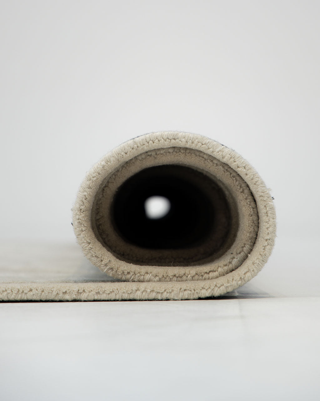 A close-up side view of the RAMSHA HOME Handmade irregular 200x300 cm Rug HTWV-191 in cream and beige, shown rolled up on a light-colored surface, with its interior forming a tunnel-like effect.
