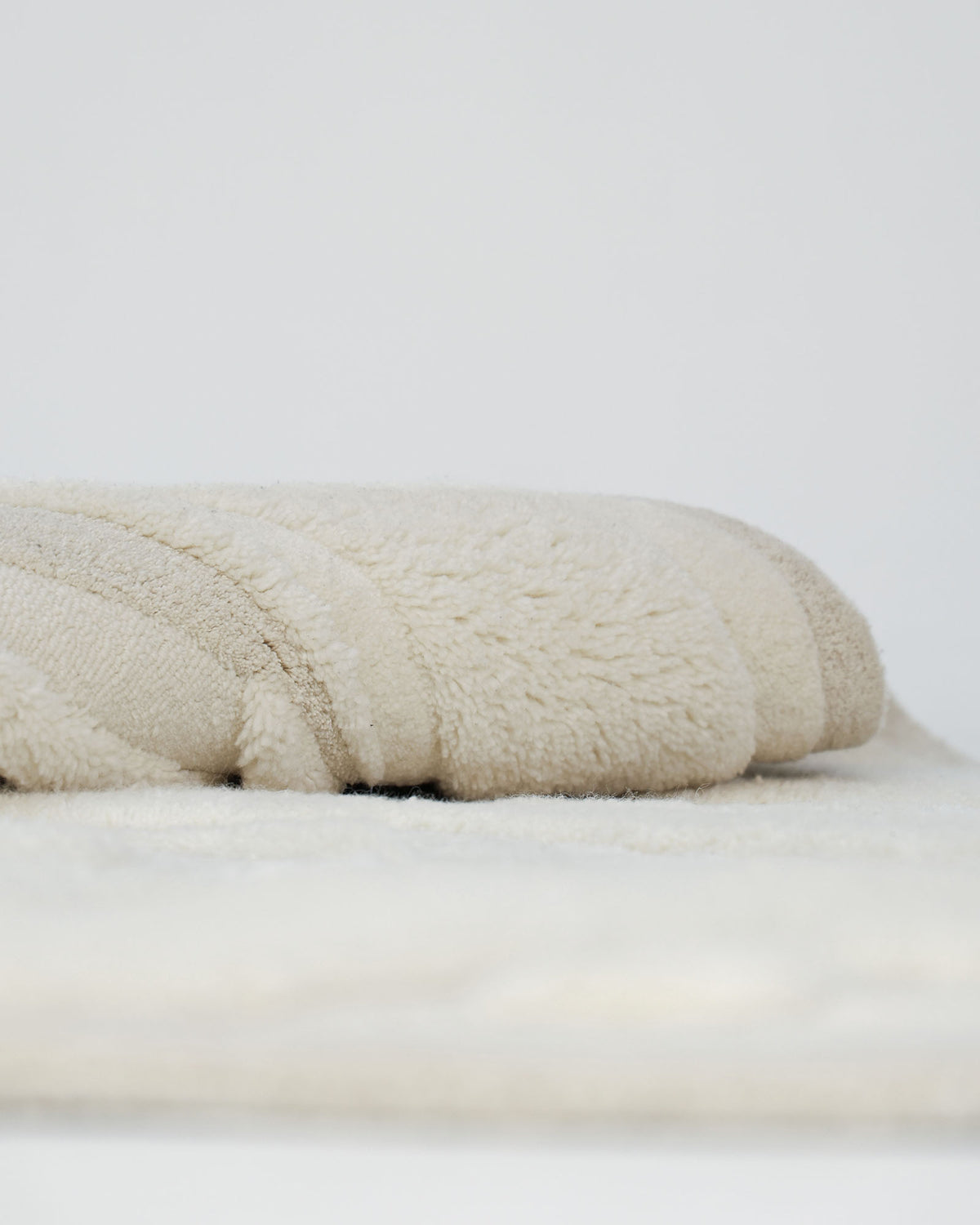 A close-up of a folded, fluffy beige towel resting on a soft white surface, beside the RAMSHA HOME Beige Color Oval Shaped Handmade Rug - HTWV 106 (200x300cm), set against a plain light background.