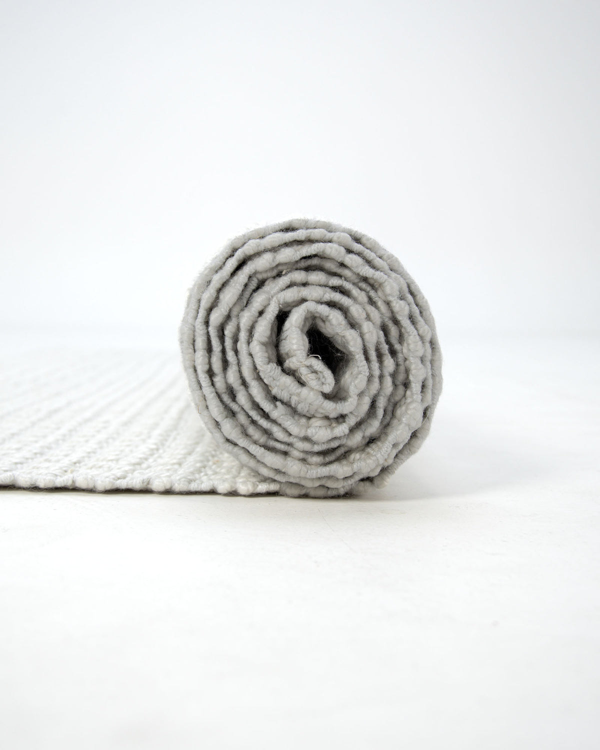 A Handmade Cream Natural Color Hand Woven Area Rug (WR-039) by RAMSHA HOME rests rolled-up on a white surface, with its textured natural fibers facing the camera and the unrolled portion extending gracefully into the background—an elegant artisanal home décor piece that infuses any space with subtle, handmade charm.