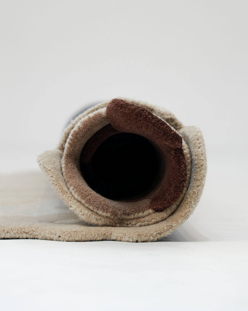 A close-up view of the RAMSHA HOME Handmade Shape Brown Color Area Rug - HTW 86, rolled up on a white surface to showcase its beige and brown handtufted wool layers and visible textures.