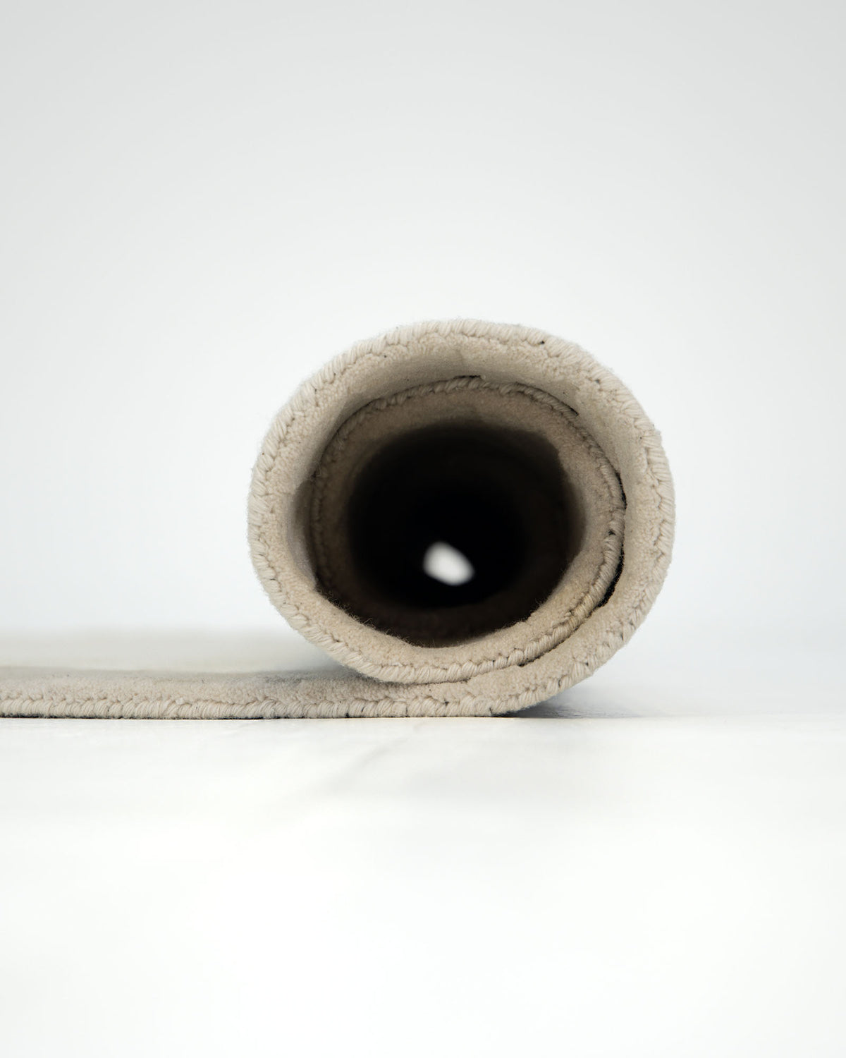 A RAMSHA HOME Beige Handmade Round Rug HTW 107 (200cm) is tightly rolled on a smooth, white surface with its hollow center and rolled edge facing the camera, beautifully highlighting its soft, textured natural fibers and expert artisanal craftsmanship.