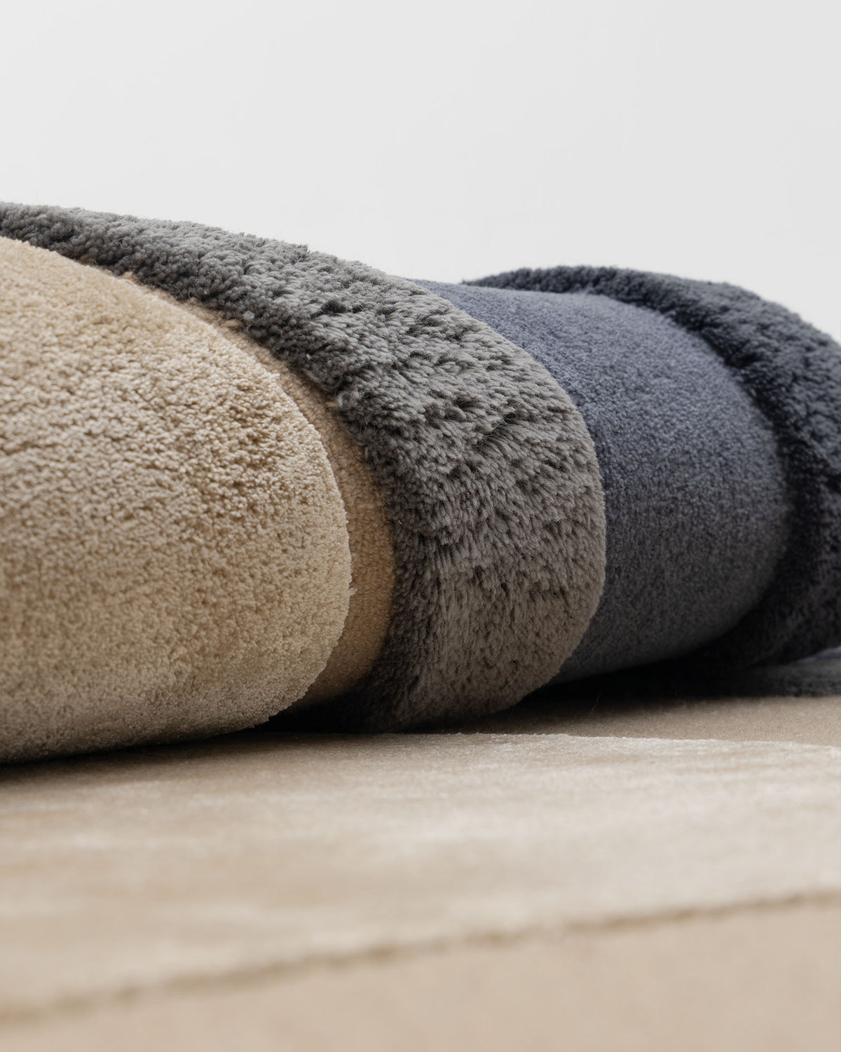 A close-up of the RAMSHA HOME Oval Shape Home Decor Multi Color Rug HTWV-90, featuring a plush modern wool texture with three visible stripes in beige, gray, and dark blue, rolled up on a beige floor against a plain white background.