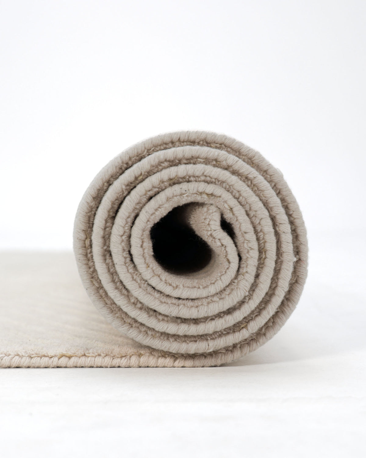 A RAMSHA HOME Home Decor Handmade Area Rug (LKH-014, 200x300 cm) in beige with a textured Scandinavian design is rolled up on a white surface, viewed from the side to display the circular layers of the rug.