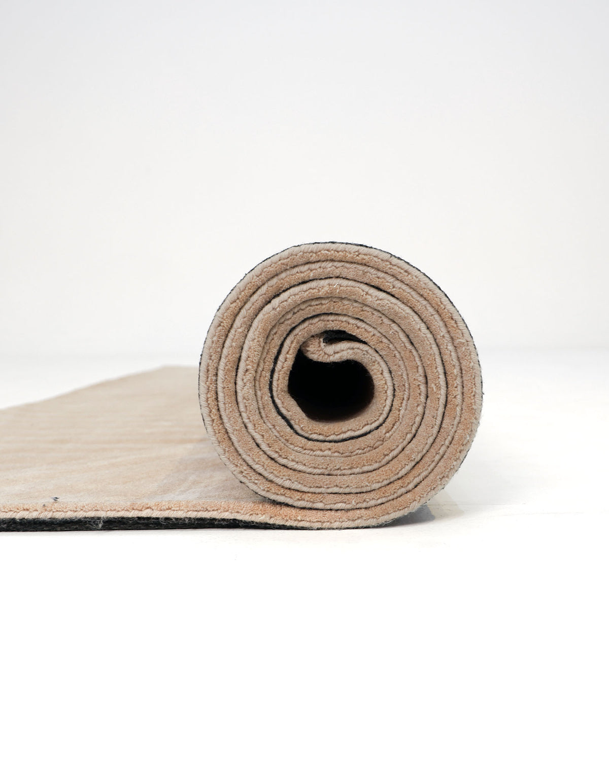 A Beige Hand Tufted Area Rug 250X350 cm For Home Decor AL-05 by RAMSHA HOME is partially unrolled and placed on a white surface, with the rolled portion facing forward and the flat end extending toward the back. This elegant rug offers subtle sophistication, making it an ideal addition to any space.