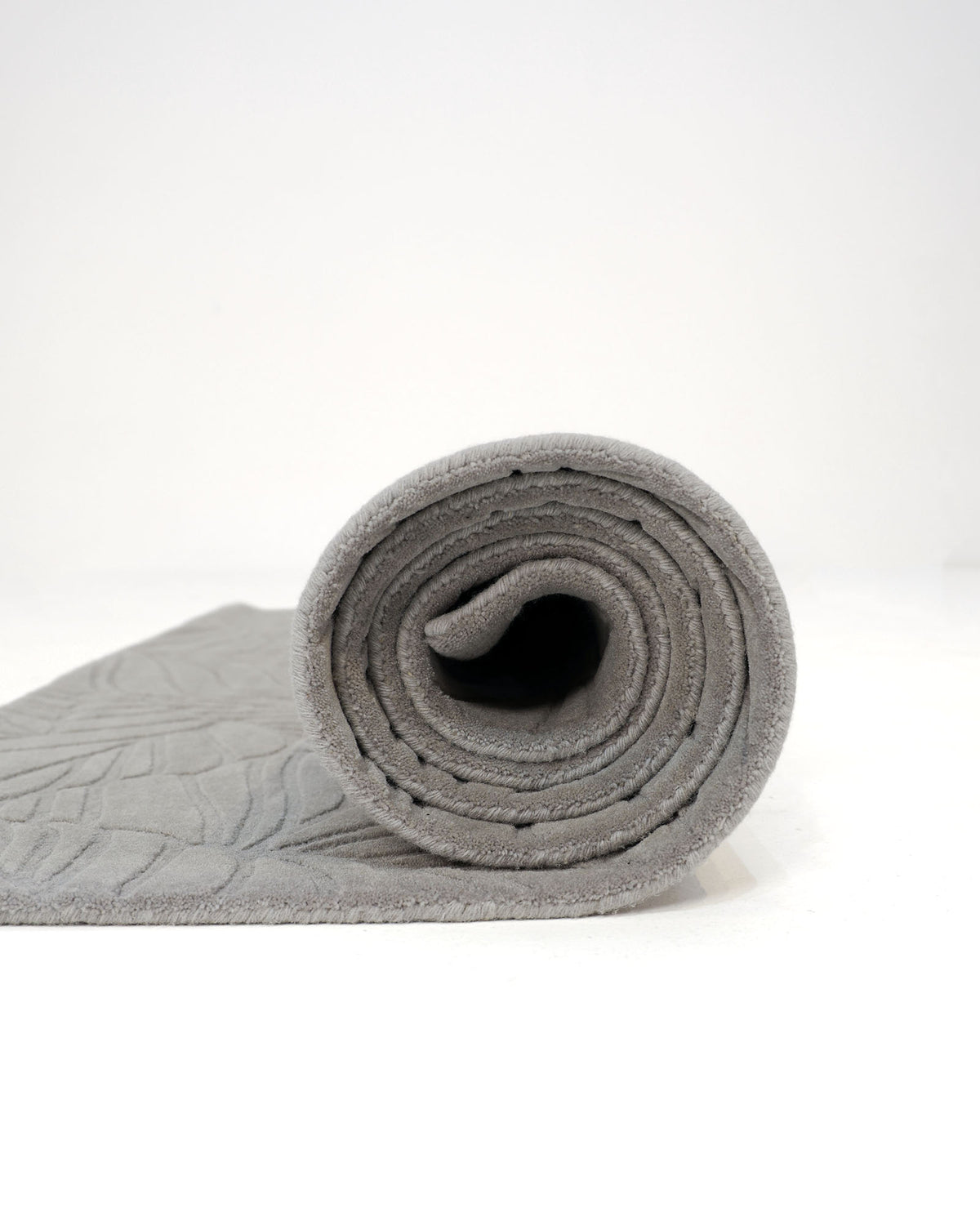 A rolled-up Rectangle Handmade Decorative Grey Color Area Rug (LKH-030) by RAMSHA HOME is displayed on a white background, showcasing its textured pattern along the surface with the roll positioned to face forward, evoking a modern neutral aesthetic.