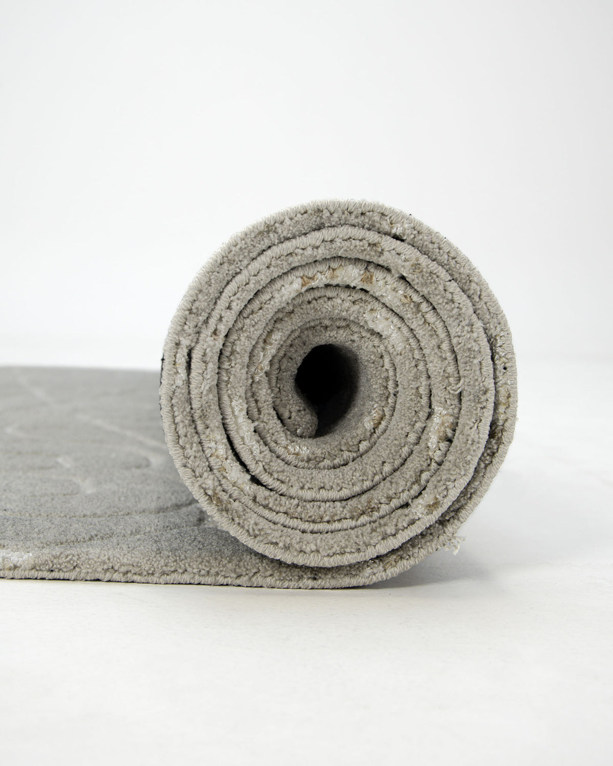 A close-up of the RAMSHA HOME Home Decor Area Rug HTP-17, crafted from light grey polyester, shown rolled up and standing upright on a white surface, with part of the transitional area rug unrolled and laying flat.