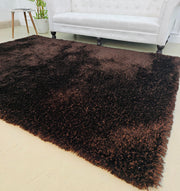 The RAMSHA HOME Fluffy Shaggy Area Rug SG 04 in brown adds warmth to a light floor in front of a white tufted sofa with dark wooden legs, with a potted plant visible in the background.
