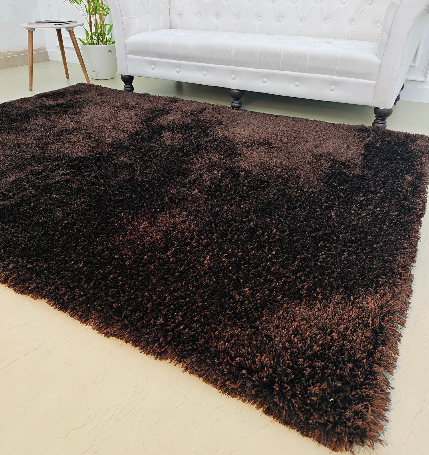 The RAMSHA HOME Fluffy Shaggy Area Rug SG 04 in brown adds warmth to a light floor in front of a white tufted sofa with dark wooden legs, with a potted plant visible in the background.