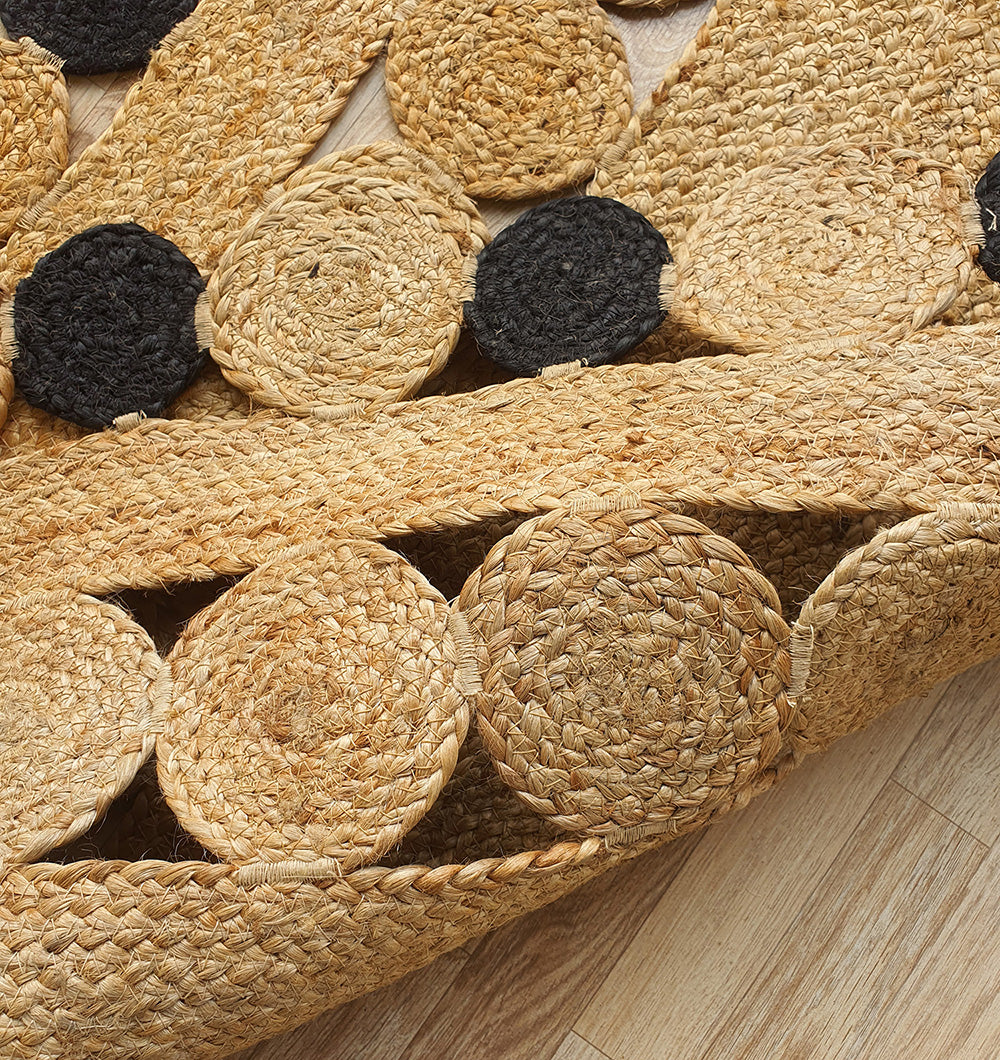 A close-up of the RAMSHA HOME Oval Shaped Jute Braided Rug Black & Natural BR-033, featuring intricate weaving and circular patterns in tan and black, displayed on a wooden floor. The natural jute texture is clearly visible.