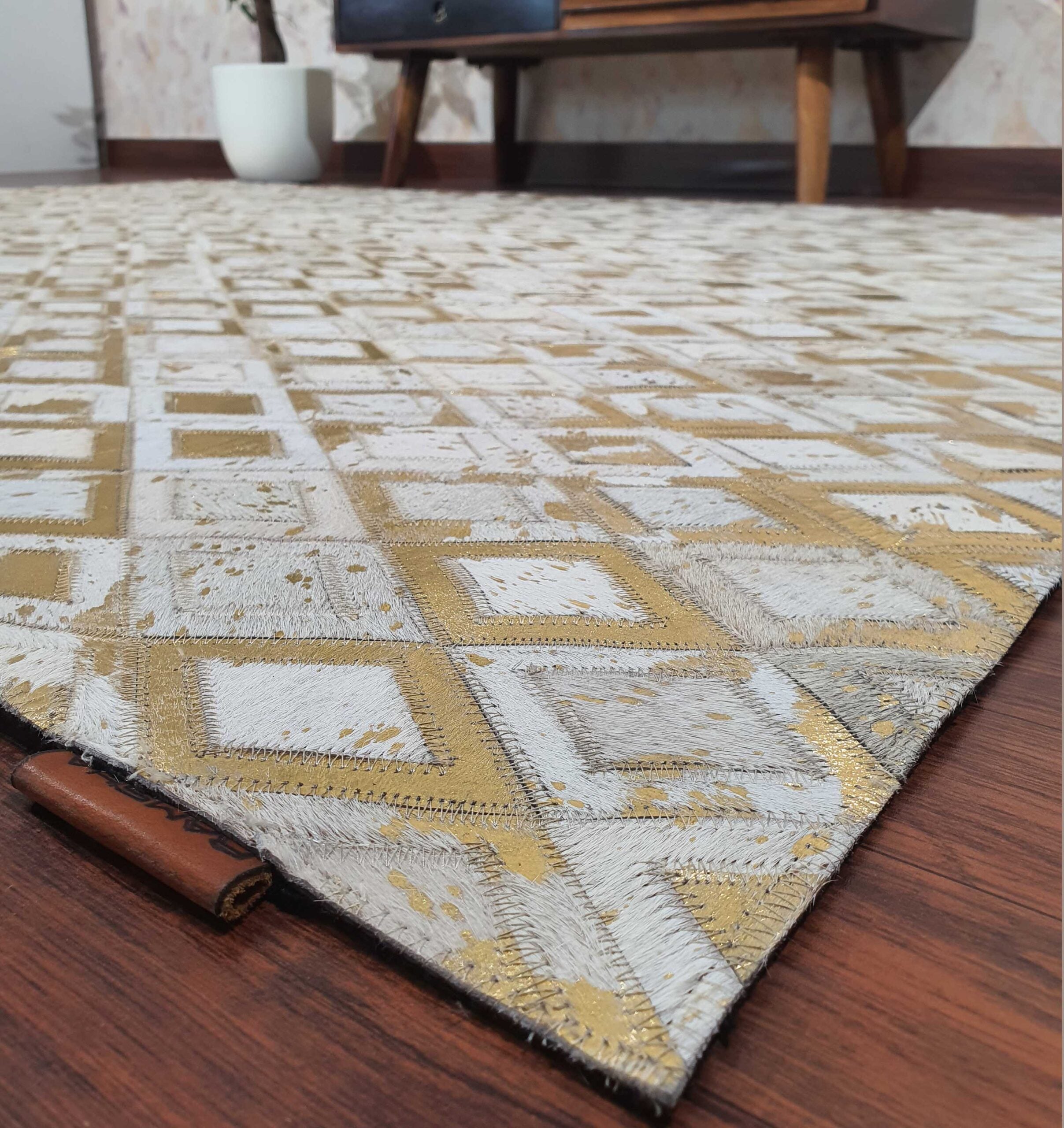 A close-up of the RAMSHA HOME Handmade Diamond Patterned Leather Rug LR-010, featuring gold and white diamond shapes on a wooden floor, with a modern wooden table and white plant pot in the background.