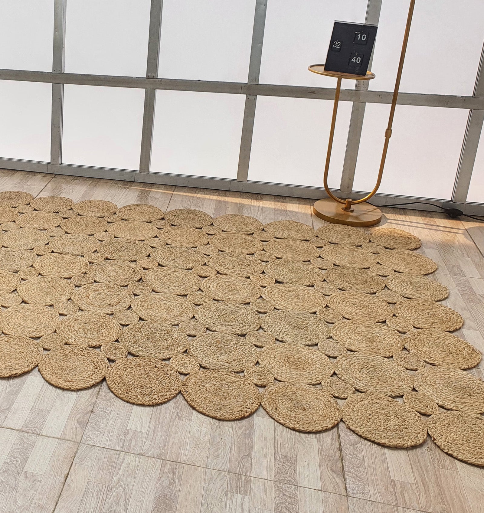 The Handmade Circular Pattern Jute Rug BR-035 by RAMSHA HOME, in beige, sits on a light wood floor next to a modern gold side table with a black block calendar. Frosted glass panels with metal frames complete the contemporary setting.