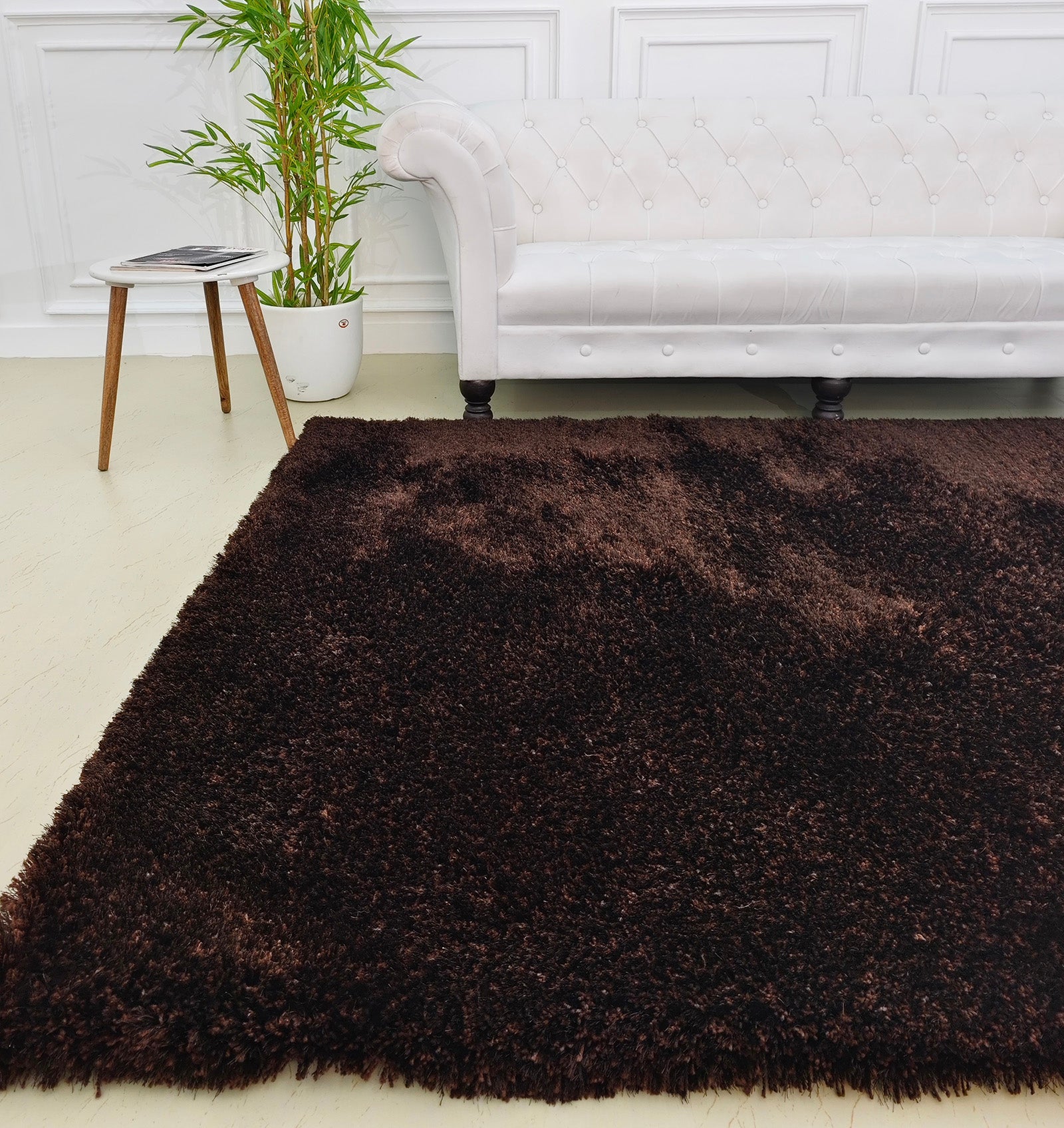 A living room with a white tufted sofa, wooden side table holding a magazine, potted plant, and the RAMSHA HOME Fluffy Shaggy Area Rug SG 04 - Brown Rectangle adding contrast to the light-colored floor.