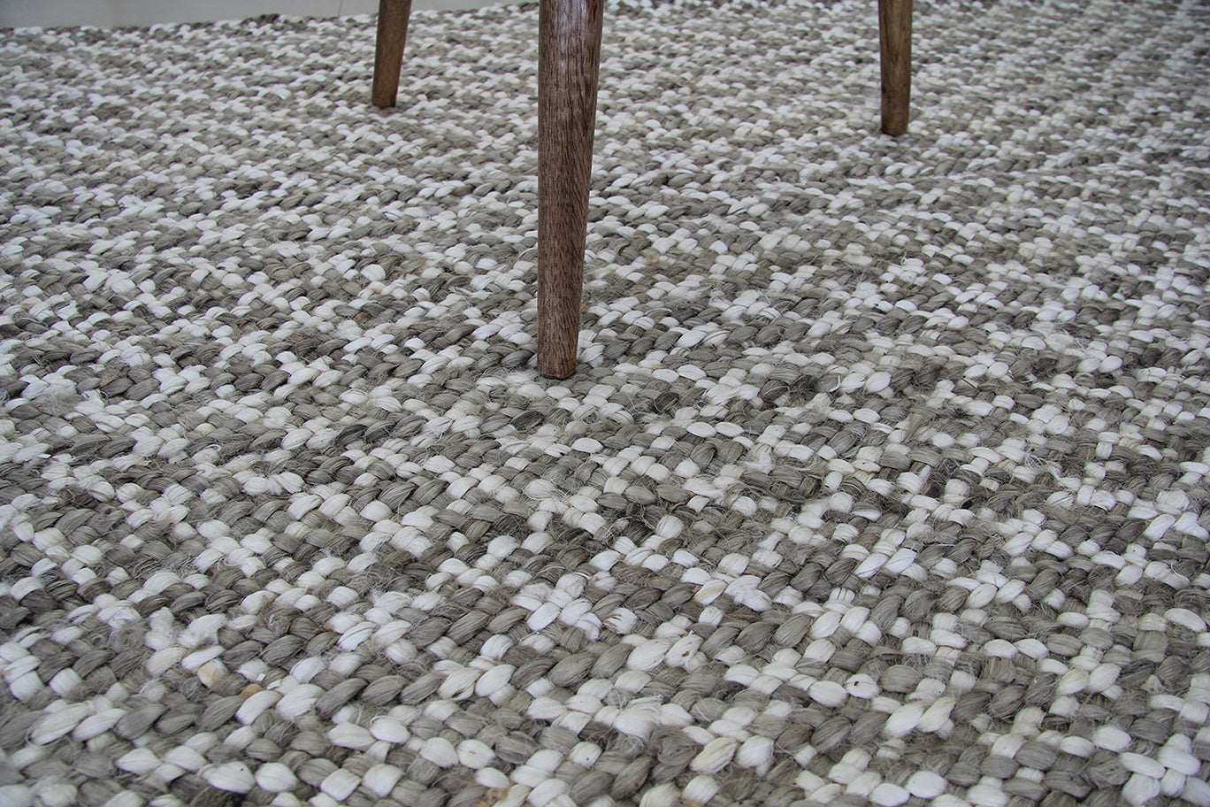 A close-up of the RAMSHA HOME Handmade Home Decor Crochet Rug - CR 003 (200x300cm) in gray, white, and cream shows its textured pattern, with three wooden legs of a chair or table visible at the top.