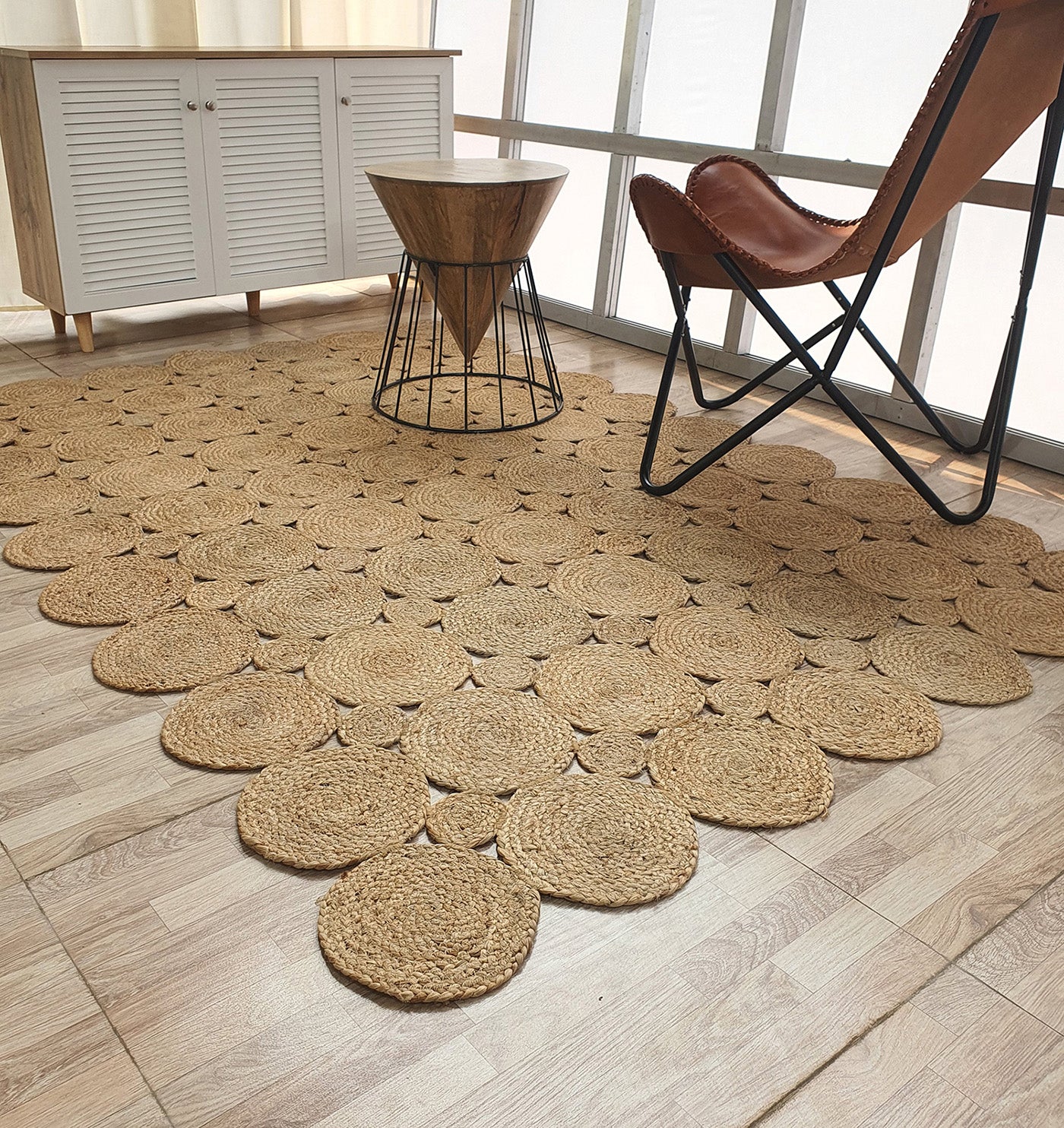 A modern room features the RAMSHA HOME Handmade Circular Pattern Jute Rug BR-035, a wooden cabinet, small round wooden table, and brown leather butterfly chair on a light wood floor by large windows.