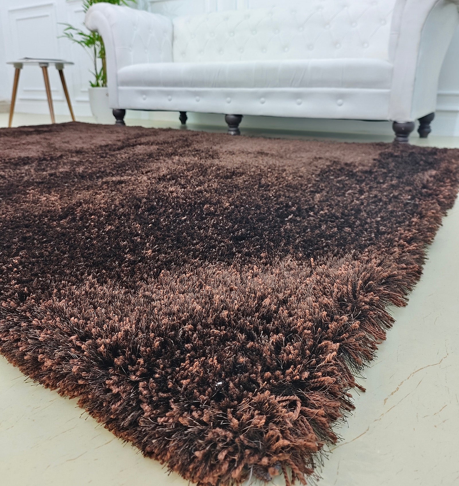 A close-up of the RAMSHA HOME Fluffy Shaggy Area Rug SG 04 in brown, featuring its plush texture on a light floor with a white tufted sofa and small side table in the background.