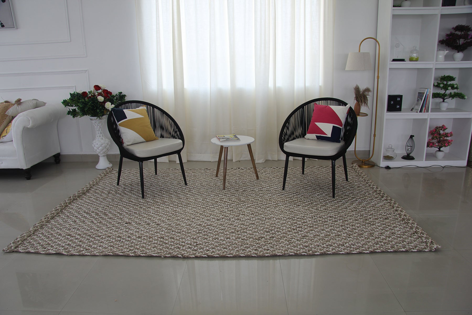 A modern living room includes two black chairs with geometric cushions, a white sofa, a round table with books, a floor lamp, decor shelves, sheer curtains, and the RAMSHA HOME Handmade Home Decor Crochet Rug - CR 003 (200x300cm).