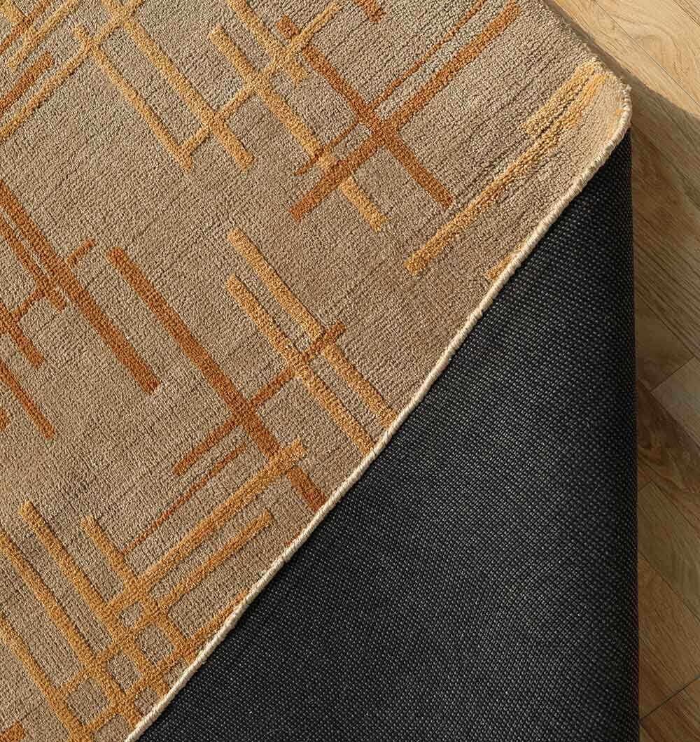A close-up of the RAMSHA HOME Luxurious Multi Color Novara Rug - CMN 34 (200x300cm), featuring a tan and orange geometric pattern, partially flipped to display its textured black non-slip backing on a light wooden floor.