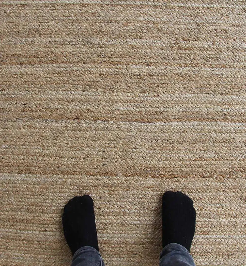A person in black socks and gray pants stands on the RAMSHA HOME Luxurious Handmade Braided Jute Floor Unshaped Rug - BR 062 (170x240cm), beige tones, viewed from above.