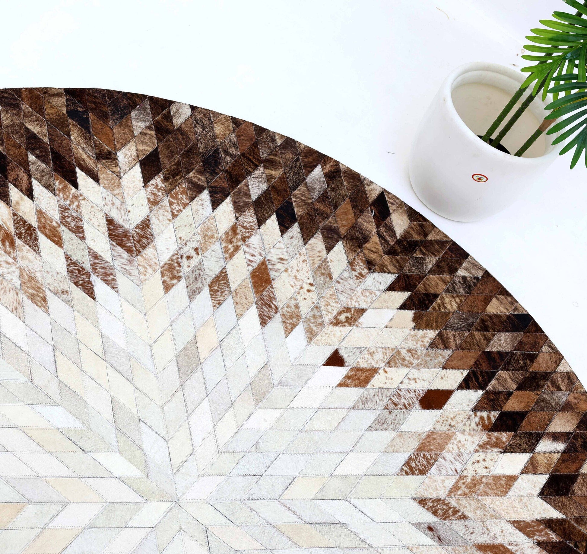 The RAMSHA HOME Multi Color Guest Room Round Decor Leather Rug - LR 022, featuring a geometric chevron pattern in brown, beige, and white, is partly shown beside a white pot with a green leafy plant on a white surface.