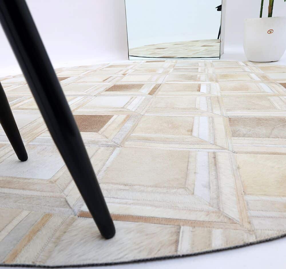 A close-up of the RAMSHA HOME Multi Color Round Leather Rug - LR 023 features geometric beige patterns, with a black table leg in the foreground and a white round planter and mirror in the background.