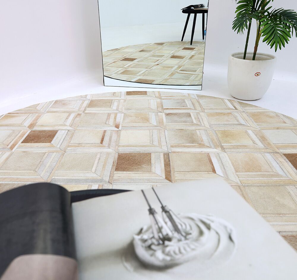 The RAMSHA HOME Multi Color Round Leather Rug - LR 023, featuring a geometric beige and tan pattern, sits on a white floor. A mirror reflects the rug and part of a black stool, with a potted plant and an open magazine nearby.