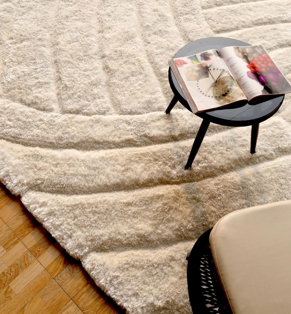A round black side table with an open magazine rests on the RAMSHA HOME Textured White Shaggy Area Rug for Large Living Room - MP3D 07 beside part of a beige chair, atop a wooden floor.