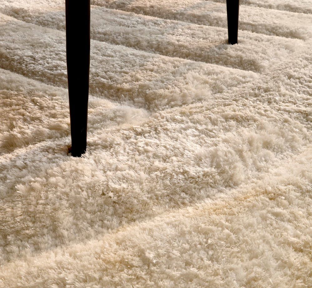 Close-up of RAMSHA HOME's Textured White Shaggy Area Rug for Large Living Room - MP3D 07, with two black furniture legs pressing into the soft ivory fibers, creating visible plush indentations and carved wave-like textures.