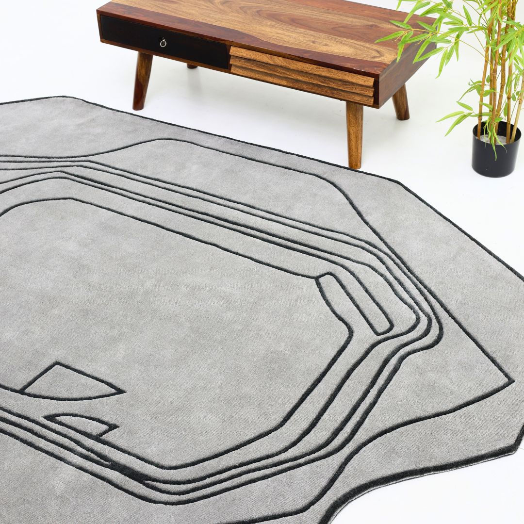 A modern living room featuring the RAMSHA HOME Shaped Rug Grey & Black Color Carpet (CMN-05) with abstract black lines, a wooden coffee table with angled legs, and a small potted plant on a white floor.